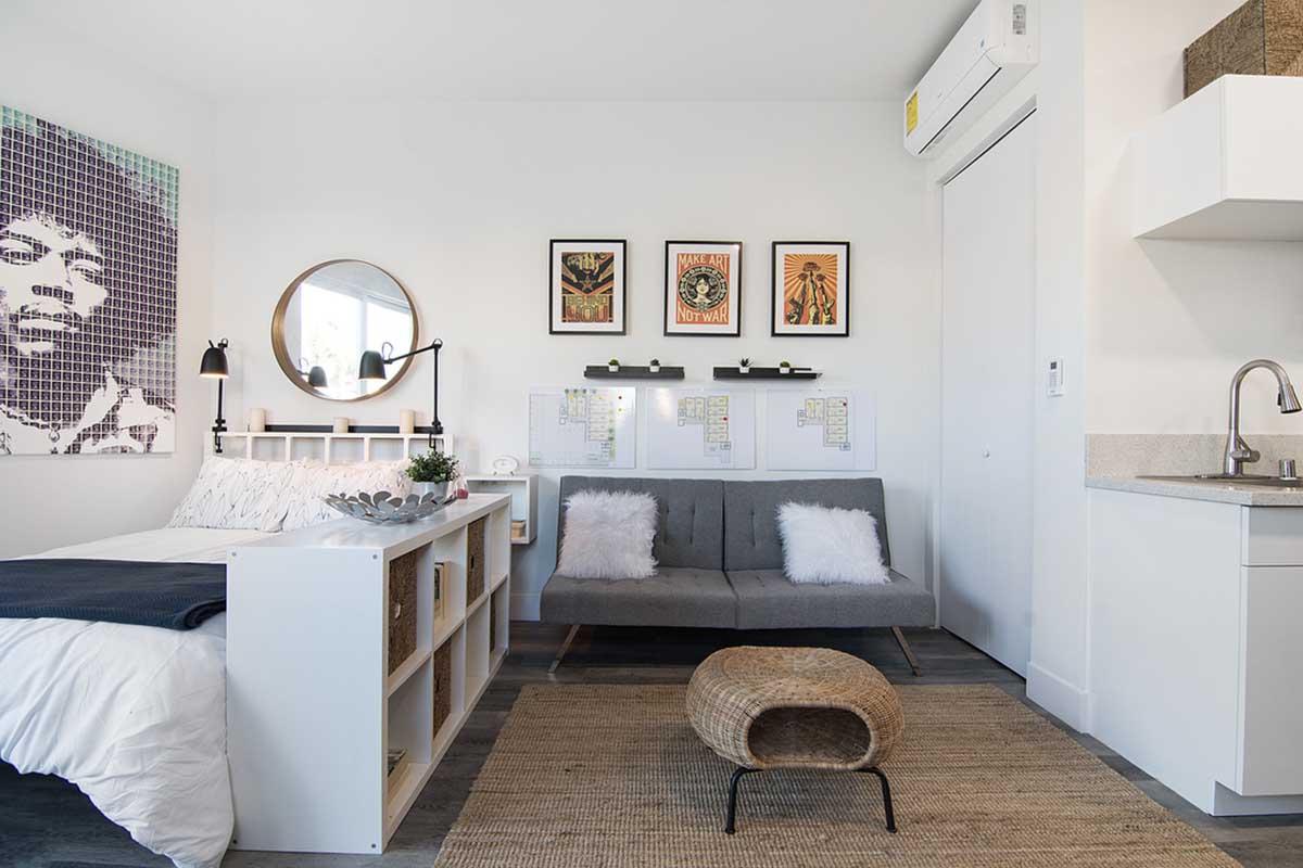 A modern, cozy bedroom featuring a bed with white linens and a decorative headboard, a gray sofa with fluffy pillows, a round mirror, and three framed posters on the wall. A small kitchen area is visible, with a kitchenette and sink, and a woven rug on the floor.