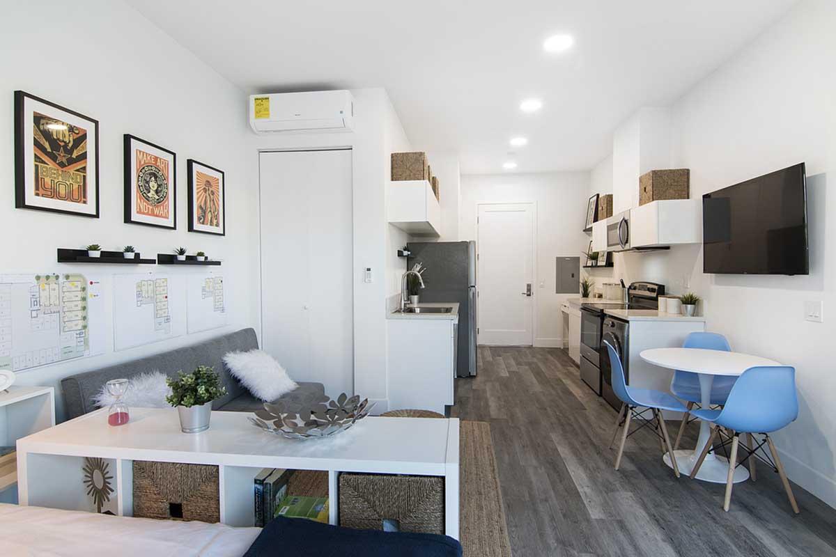 A modern studio apartment featuring a grey sofa, a small dining table with blue chairs, and a kitchen area with white cabinetry. The walls are adorned with framed artwork, and there is a television mounted on one side. The space is bright with overhead lighting and a minimalist design.