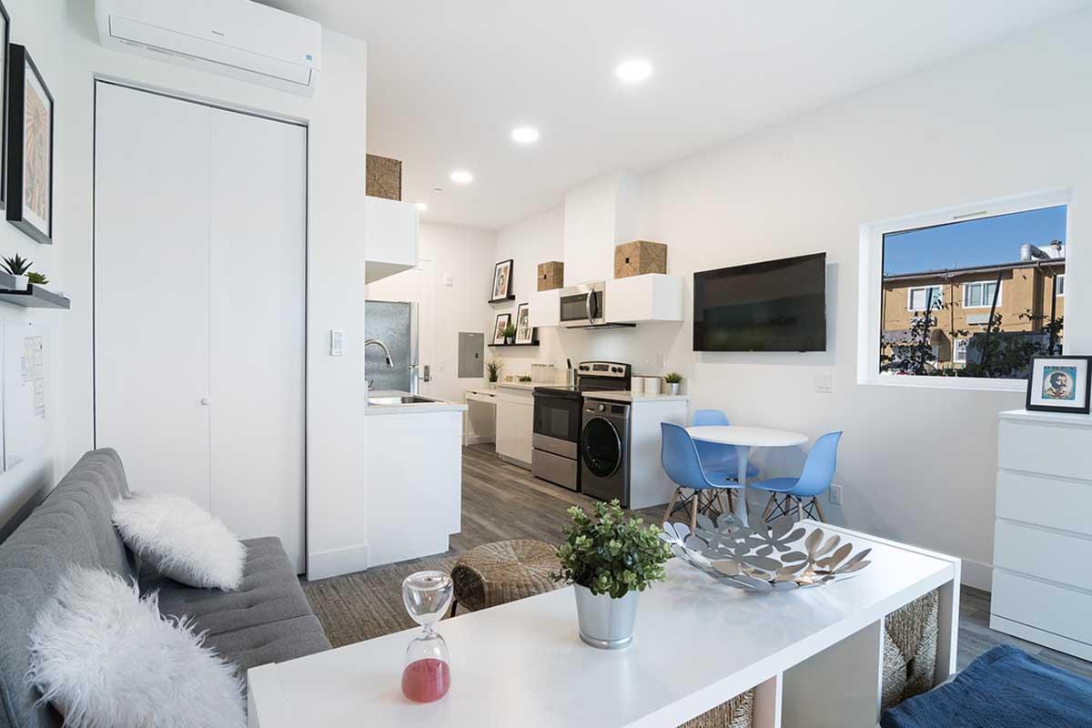 A modern, minimalist living space featuring a gray sofa with fluffy pillows, a coffee table, and a small dining area with a round table and blue chairs. The kitchen is equipped with stainless steel appliances and white cabinetry. Bright lighting and large windows create an open and airy atmosphere.