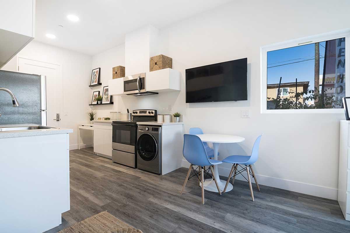 A modern kitchen featuring a compact design with a stove, microwave, and refrigerator. There's a wall-mounted TV and a small table with four blue chairs. The space is bright with natural light coming through a window, and minimalist decor enhances the contemporary feel. The flooring is light wood.