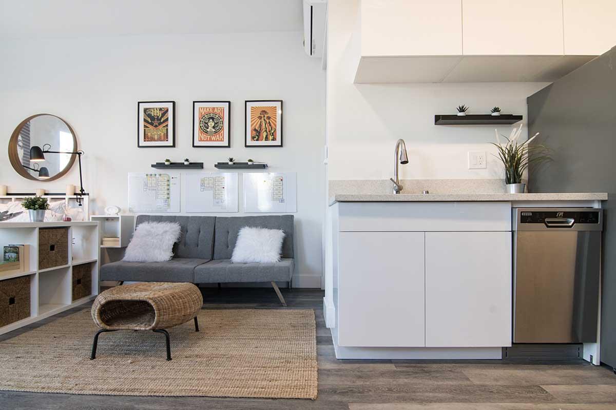 A modern, cozy living space featuring a gray sofa with white pillows, a round mirror, and framed art on the wall. To the right, a minimalist kitchen with white cabinetry, a stainless steel dishwasher, and a small table next to a woven ottoman. A natural fiber rug adds warmth to the room.