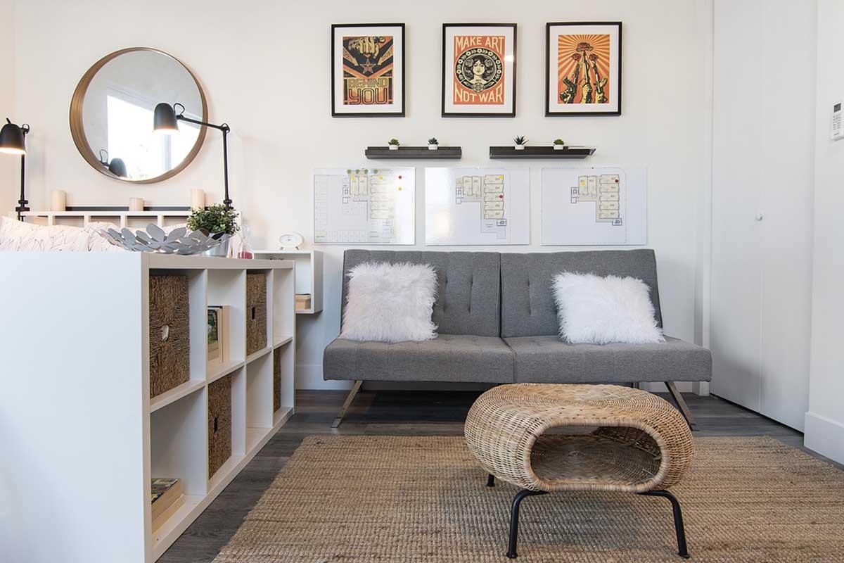 A cozy living space featuring a gray futon with white cushions, a round mirror, decorative framed posters on the wall, and a woven coffee table. There are shelves with various items and a small plant, along with a light-colored rug on the floor, creating a warm and inviting atmosphere.