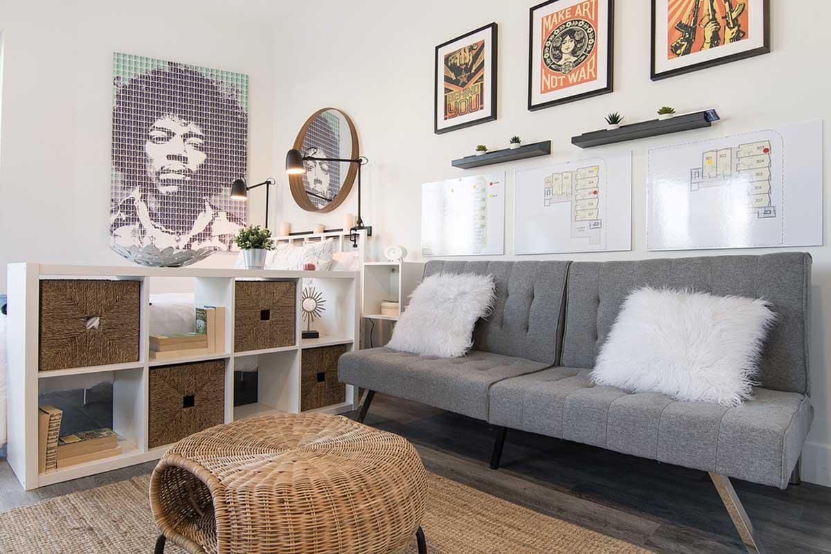 A modern living room featuring a gray sofa adorned with fluffy white pillows. Next to it, a white shelving unit with open and closed storage compartments. The wall displays framed art, including a large portrait and two smaller posters. A round decorative basket table completes the cozy, stylish space.