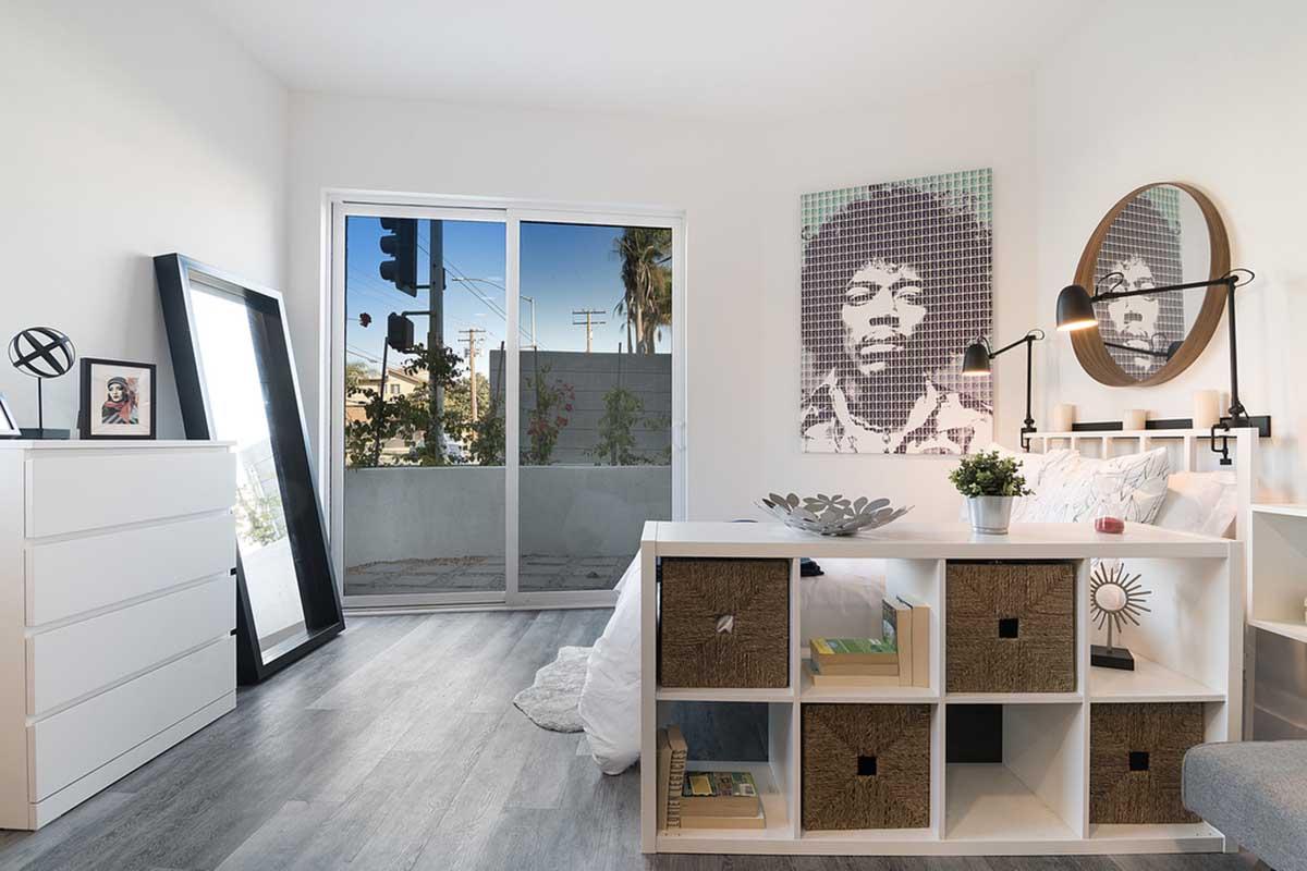 A cozy bedroom featuring a white wall with a large window, allowing natural light. There's a modern dresser with a large mirror leaning against the wall. A shelf unit with decorative storage boxes sits in front of the bed, which has white linens. A wall art piece of a musician adds personality to the room.