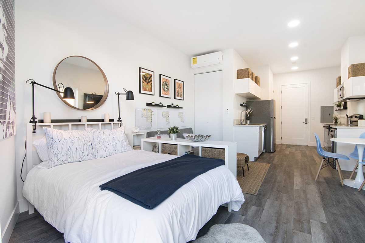 A modern studio apartment featuring a queen-sized bed with white bedding and a navy throw, a round mirror above the bed, and artwork on the walls. The space includes a small dining table, kitchenette with appliances, and stylish wooden accents, all in a bright and airy layout.