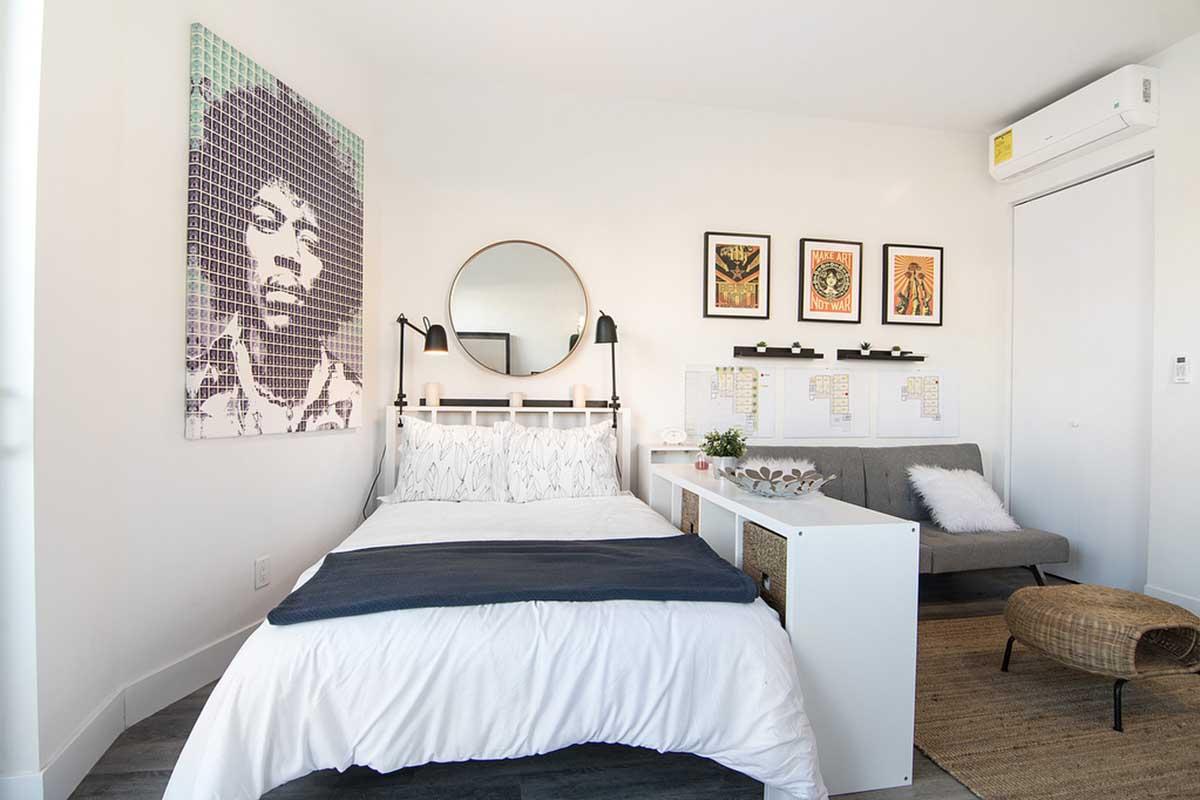 A cozy bedroom featuring a white bed with a blue throw blanket, a circular mirror above the headboard, and artwork on the wall, including a portrait of a musician. A small gray sofa is in the corner, and there are framed illustrations above a desk space. The room has light walls and modern decor elements.