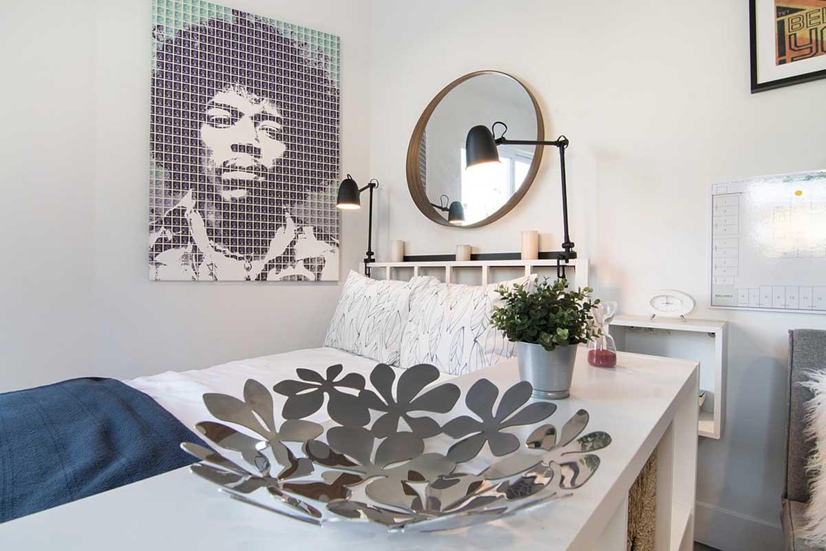 A modern bedroom featuring a bed with patterned pillows and a blue throw. Above the bed is a large, colorful mural of a musician's face. A round mirror and stylish lamps are mounted on the wall, while a silver flower-shaped bowl sits on a white tabletop. A small potted plant adds a touch of greenery to the space.