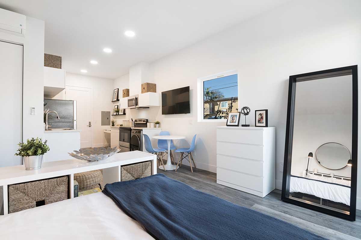 A modern studio apartment featuring a cozy bed with blue bedding, a minimalist white table with blue chairs, a large mirror, and a kitchenette. The space is bright and airy, with sunlight coming through a window. Decor includes framed pictures and decorative plants.