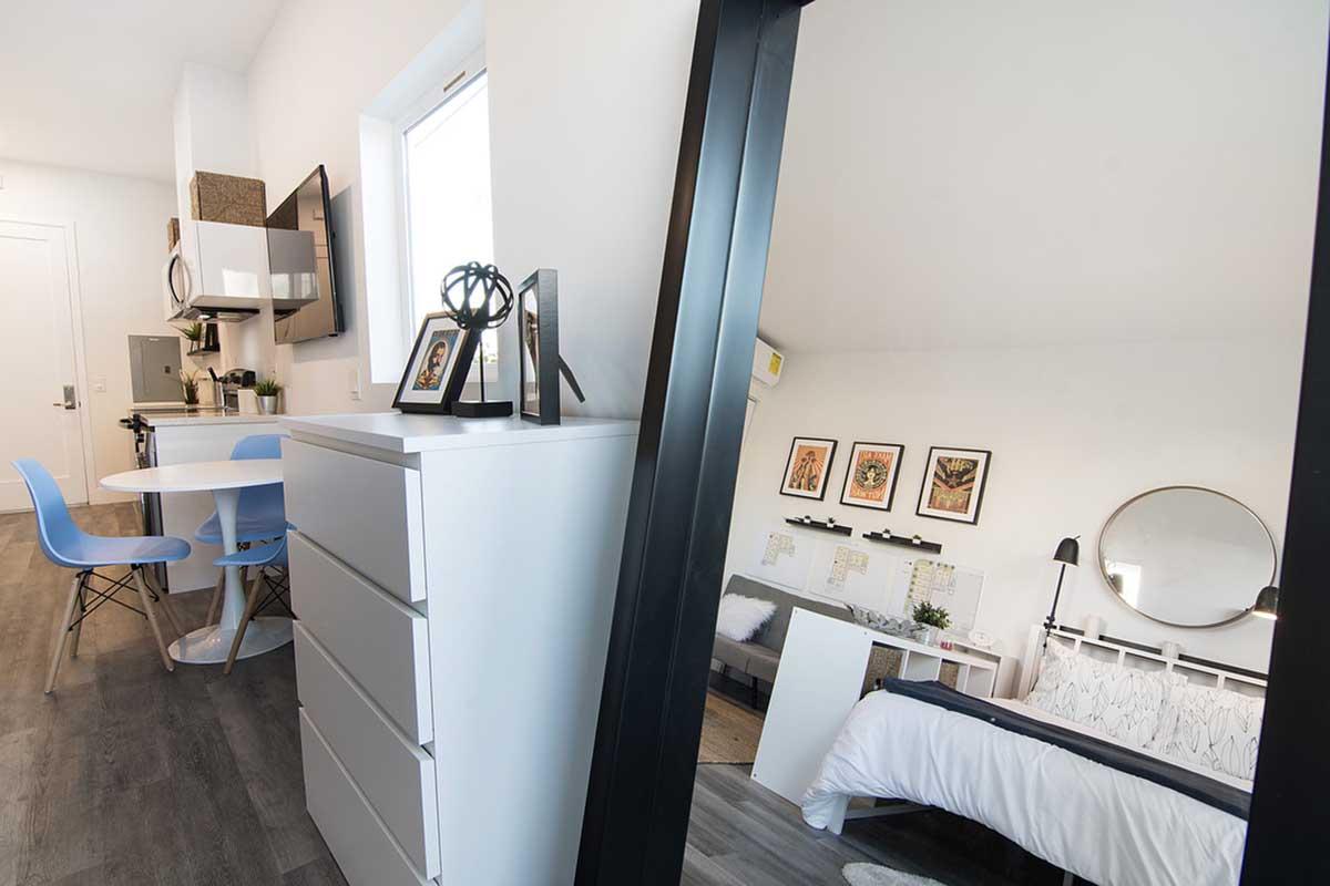 A modern, minimalist studio apartment featuring a small dining area with a round table and blue chairs, a kitchenette, and a bedroom with a bed, decorative wall art, and a large mirror reflecting the space. Natural light streams in through a window, enhancing the bright, open atmosphere.
