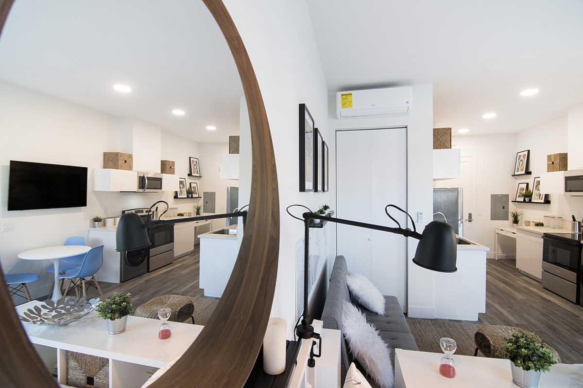 Modern kitchen and living area seen through a round mirror. The kitchen features white cabinetry, stainless steel appliances, and a dining table with blue chairs. A cozy seating area includes a dark sofa and decorative plants, creating a bright and inviting atmosphere.