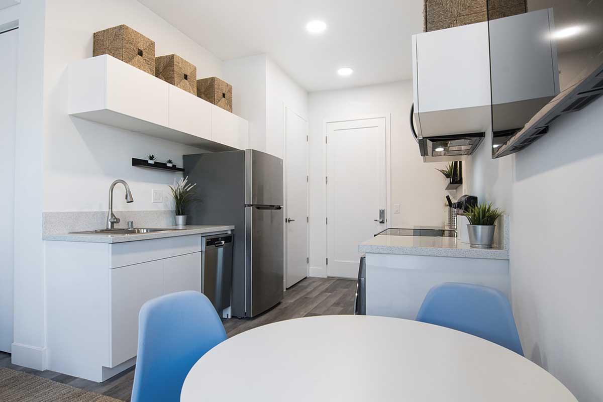 Modern kitchen featuring white cabinetry, a stainless steel refrigerator, and a simple round dining table with blue chairs. Storage bins are placed on the shelves, and there are plants for decoration. The space is bright and minimalistic, with sleek appliances and a light color palette.