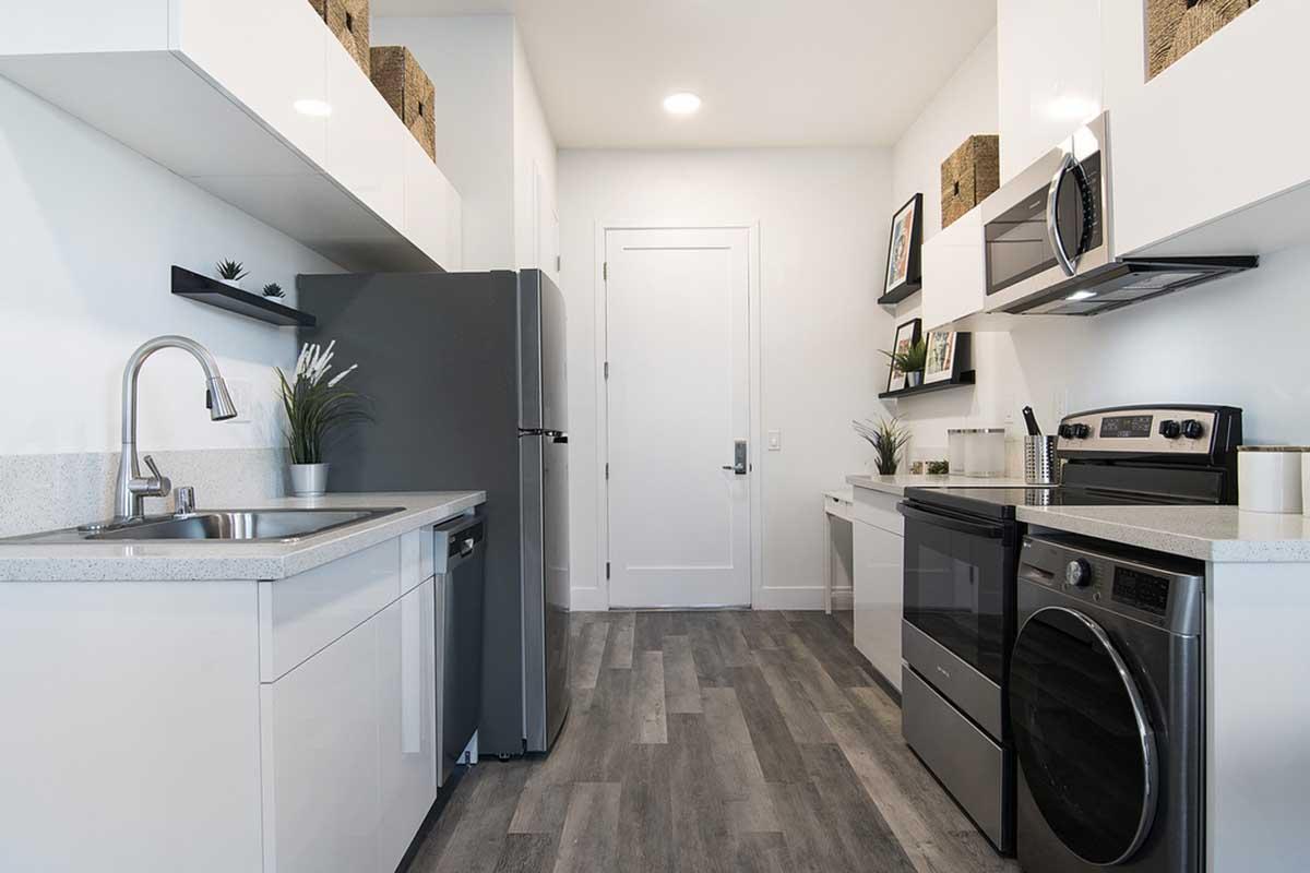 A modern kitchen featuring sleek appliances, including a refrigerator, oven, and washer/dryer combo. The design includes white cabinets, light countertops, and decorative baskets on shelves. Natural light illuminates the space, which has a minimalist aesthetic with plants and neutral tones.