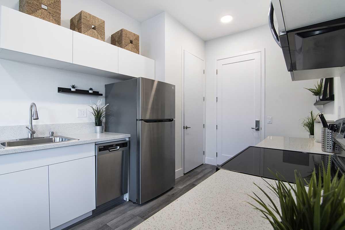 Modern kitchen featuring stainless steel appliances, including a refrigerator and stove. The countertop is sleek and white, complemented by a sink and a dishwasher. Above, there are minimalist cabinets and decorative storage baskets. The walls are light-colored, and there are small plants for a touch of greenery.
