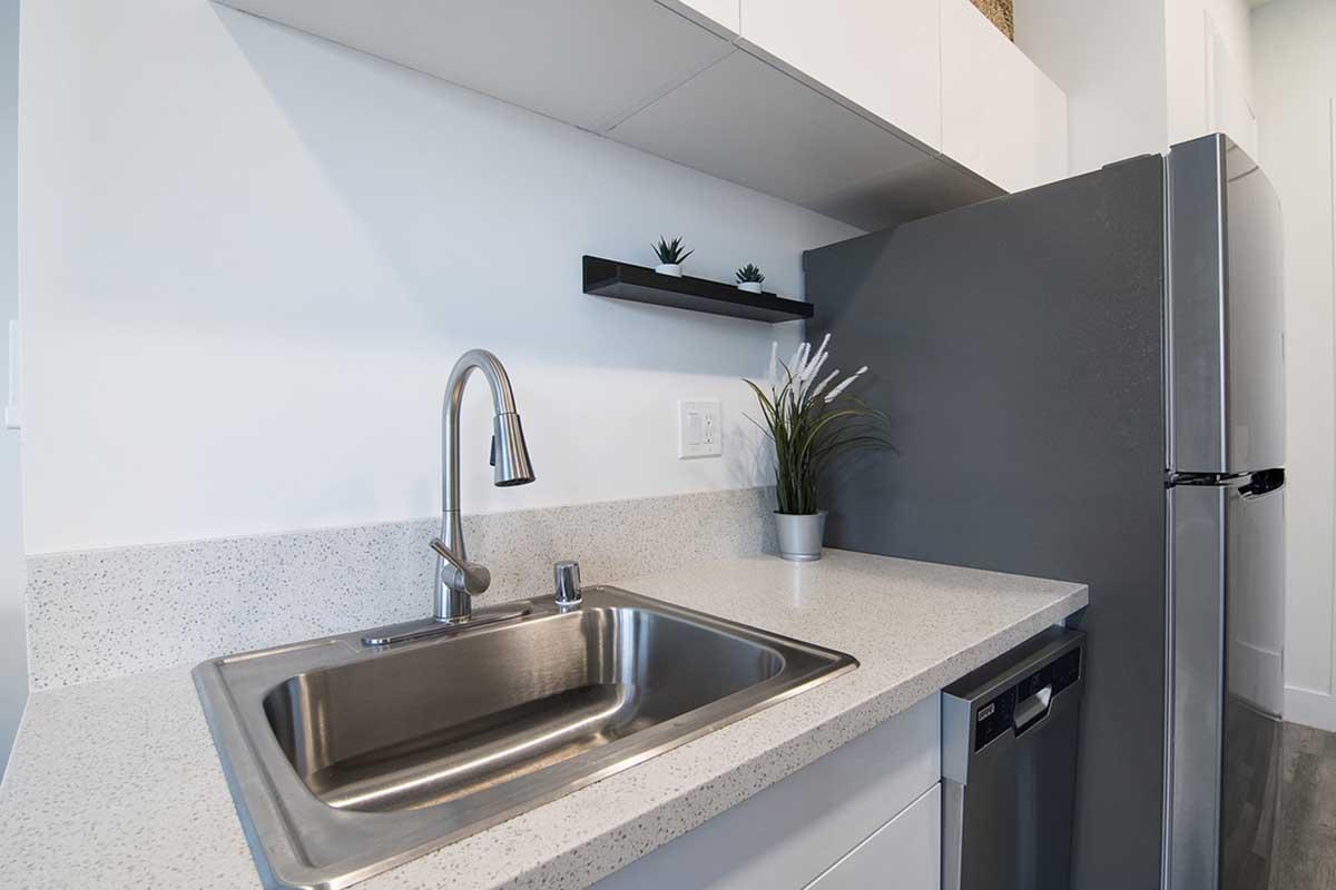 A modern kitchen scene featuring a stainless steel sink with a sleek faucet, a gray refrigerator, and minimalist white cabinetry. A small plant sits on the countertop alongside a potted arrangement, and a shelf above holds decorative items. The design emphasizes clean lines and a contemporary aesthetic.
