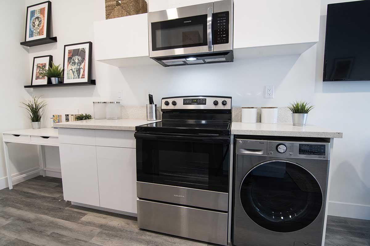 A modern kitchen layout featuring a black stove, silver microwave above it, and a front-loading washer. The countertop is light-colored with decorative plants and framed artwork on the wall. There's a small workspace area with additional storage beneath, creating a functional and stylish space.