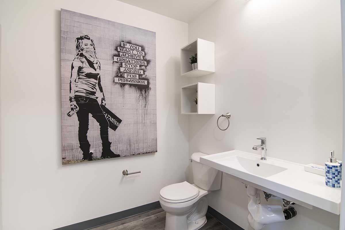 A modern bathroom featuring a minimalist design. The space includes a white sink and toilet, and there's a large, graffiti-style artwork on the wall depicting a female figure holding a spray can with the text "If you want to achieve greatness, stop asking for permission." The decor is clean and contemporary.