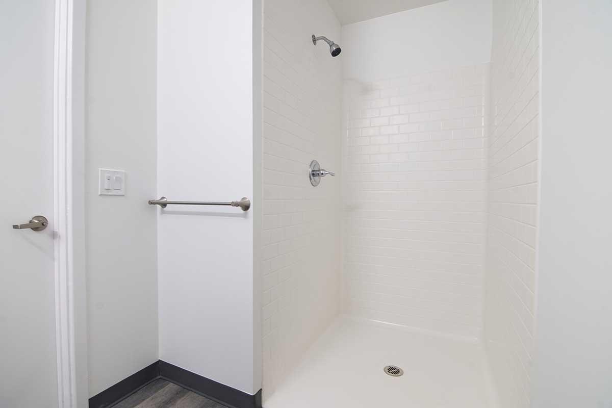 Empty walk-in shower featuring white tiled walls and a smooth floor. The space includes a chrome showerhead mounted on the wall and a grab bar. The surrounding walls are painted in a light color, enhancing the bright and airy feel of the bathroom.