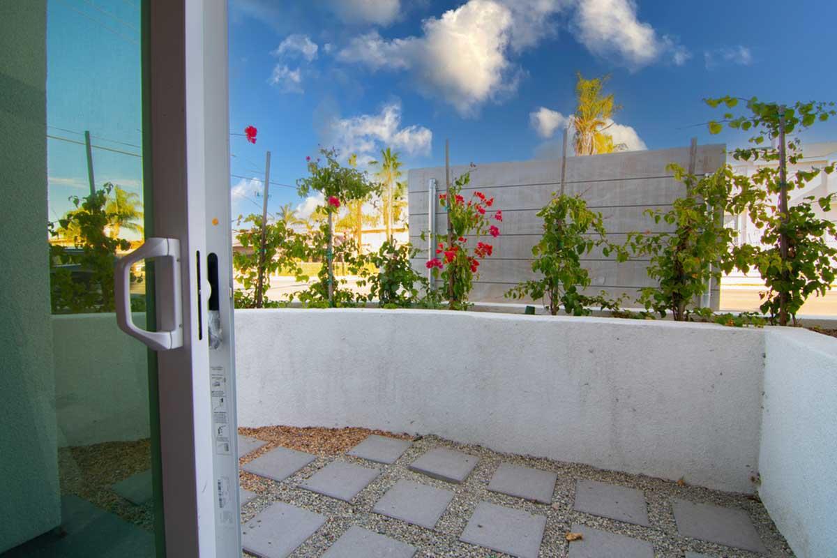 View from a glass door opening to a small patio area with square stone pavers and gravel. Lush green plants with vibrant flowers are growing along a white wall. The background features a clear blue sky with scattered clouds, creating a serene outdoor atmosphere.