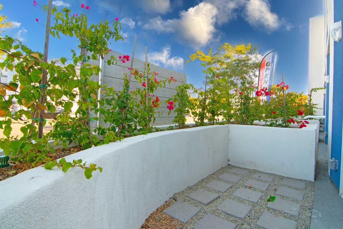 A landscaped area featuring a curved white wall with gray stone paving and flowering plants. Green foliage and vibrant flowers add color, while a flag reading "OPEN" is visible in the background against a bright sky with scattered clouds.