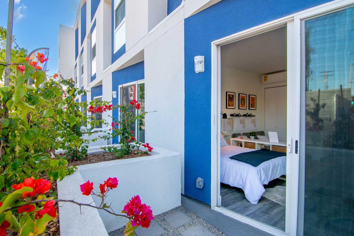A bright, inviting room opens onto a patio with vibrant pink flowers. The bed, dressed in white, is positioned near a minimalist desk. The exterior features blue and white walls, with large windows offering natural light. The scene conveys a modern, comfortable living space with a touch of nature.