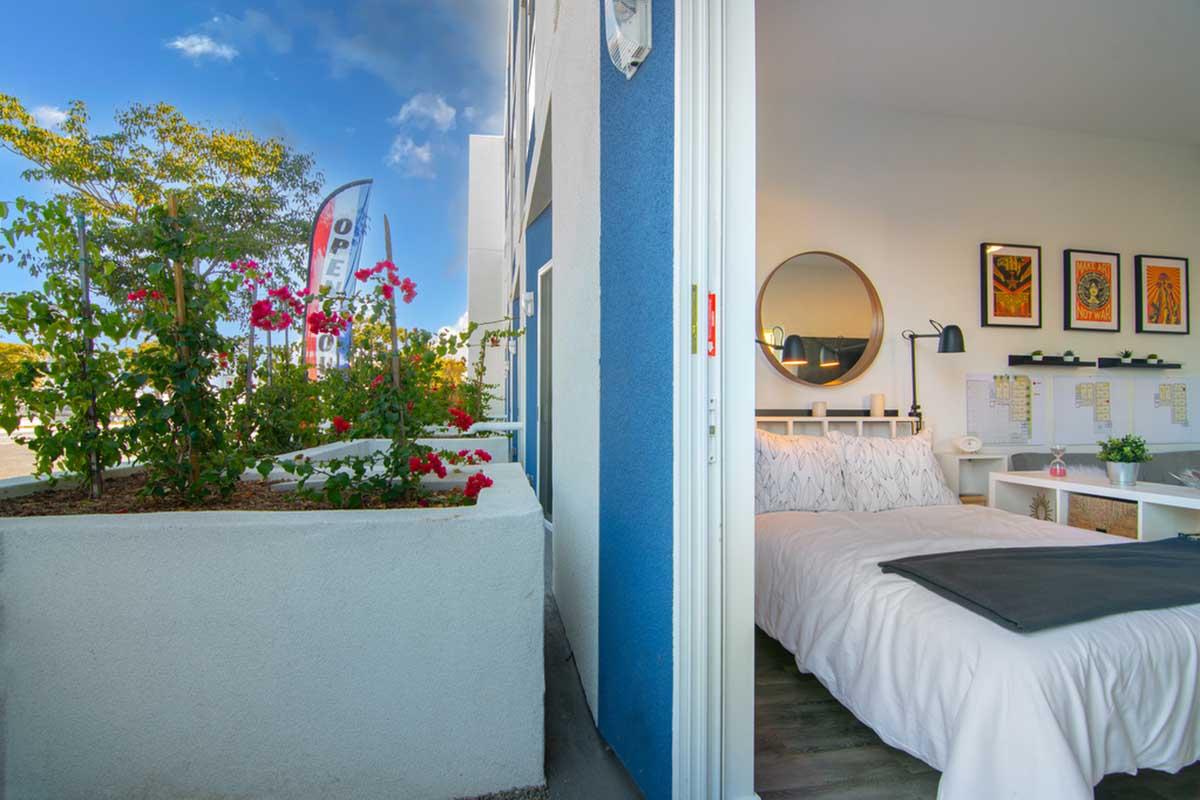 View of a bright, modern hotel room with a bed and decorative wall art. A door opens to an outdoor area featuring vibrant flowers. In the background, flags indicate the hotel is open. The room has a minimalist design with a round mirror and a desk space, blending indoor and outdoor elements.