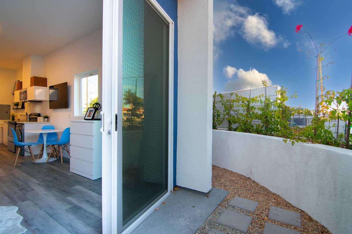A view from a modern balcony with sliding glass doors, featuring a small outdoor space with gravel and plants. In the background, a bright blue sky with clouds and a glimpse of a kitchen and dining area through the open door.