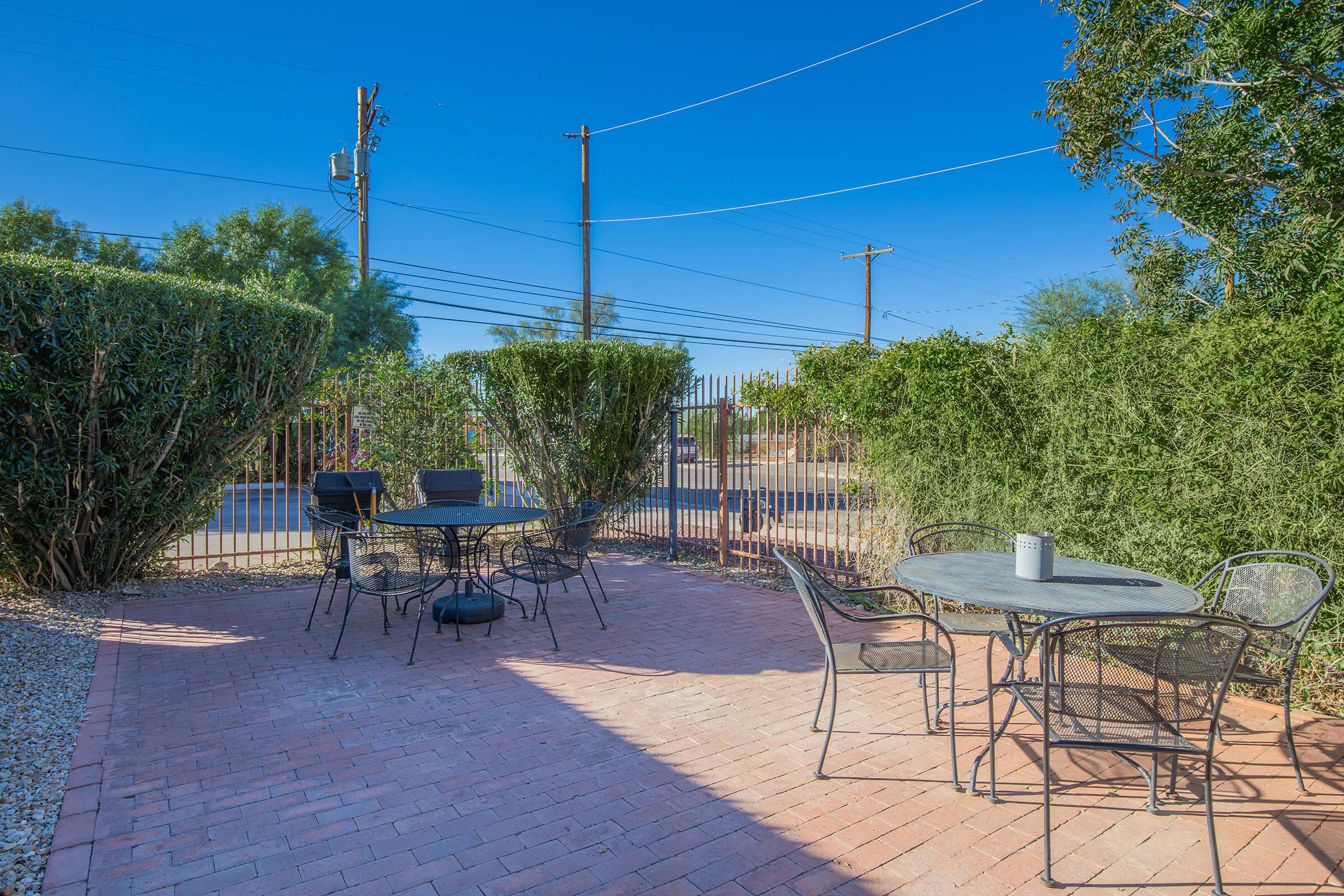 A cozy outdoor seating area featuring two round tables with chairs surrounded by green shrubs. The patio is made of brick, with a clear blue sky overhead. Nearby power lines are visible in the background, creating a peaceful yet urban atmosphere.