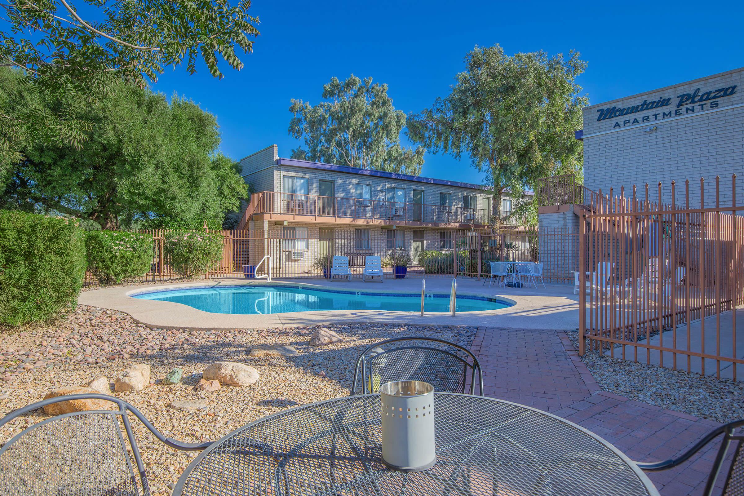 A bright outdoor scene featuring a swimming pool surrounded by a landscaped patio area at the Mountain Plaza Apartments. There are lounge chairs near the pool, a table with a candle holder in the foreground, and well-maintained shrubs and trees providing greenery.