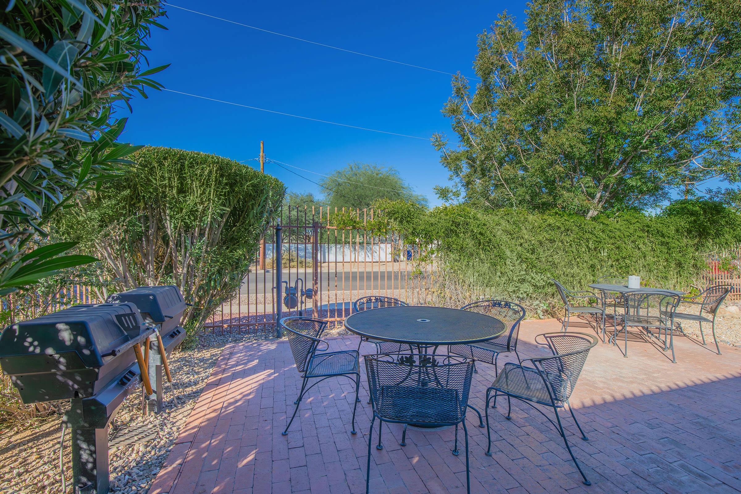 Outdoor dining area featuring a round table with several black metal chairs, surrounded by lush greenery. A grill is positioned nearby, set against a clear blue sky. The scene offers a peaceful atmosphere, ideal for relaxation or social gatherings.