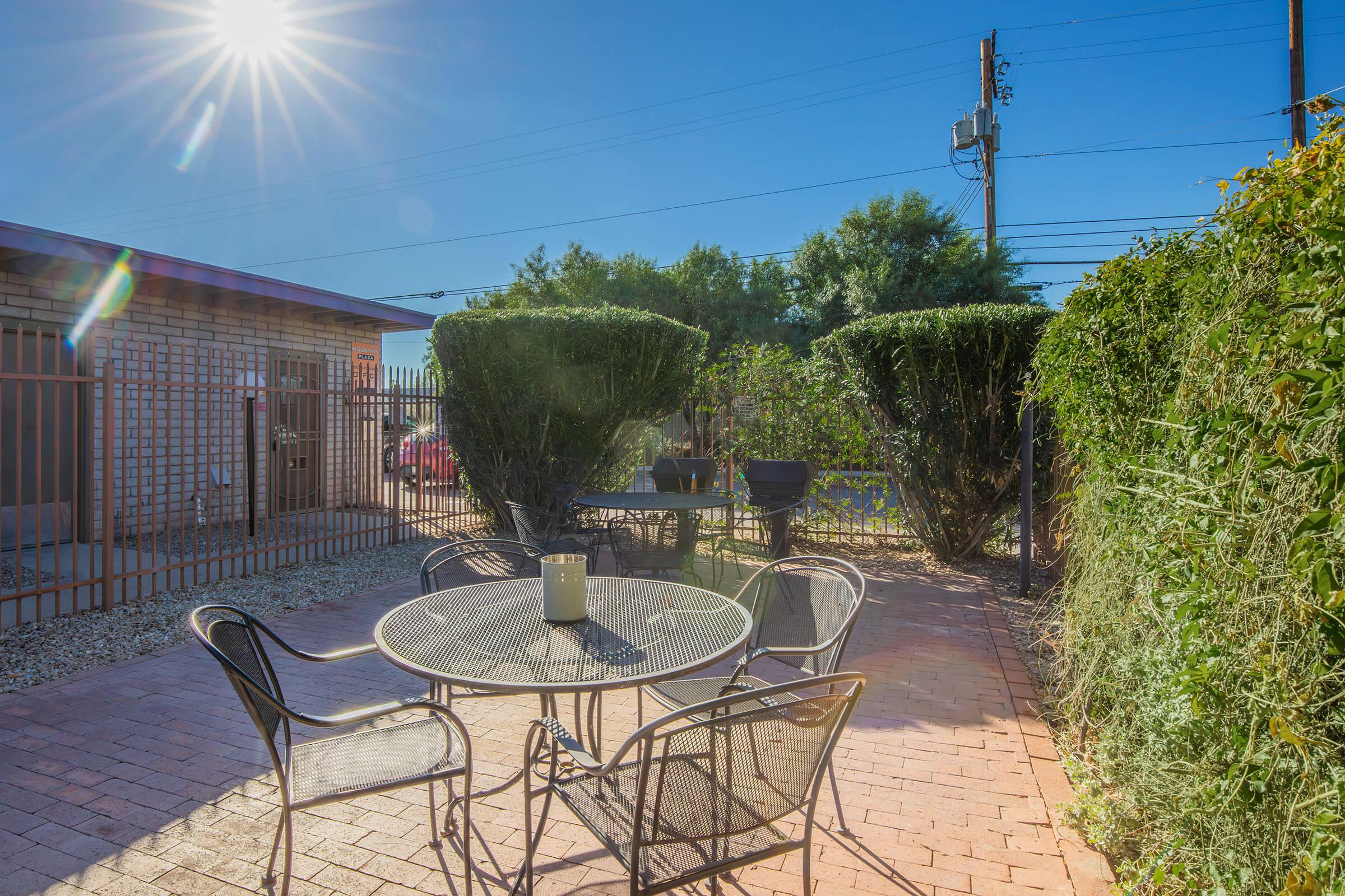 A bright, sunny backyard featuring a circular glass table surrounded by four black metal chairs. Lush green bushes and a wooden fence create a cozy atmosphere, with a grill in the background and clear blue skies overhead. The scene conveys a serene outdoor space perfect for relaxation.