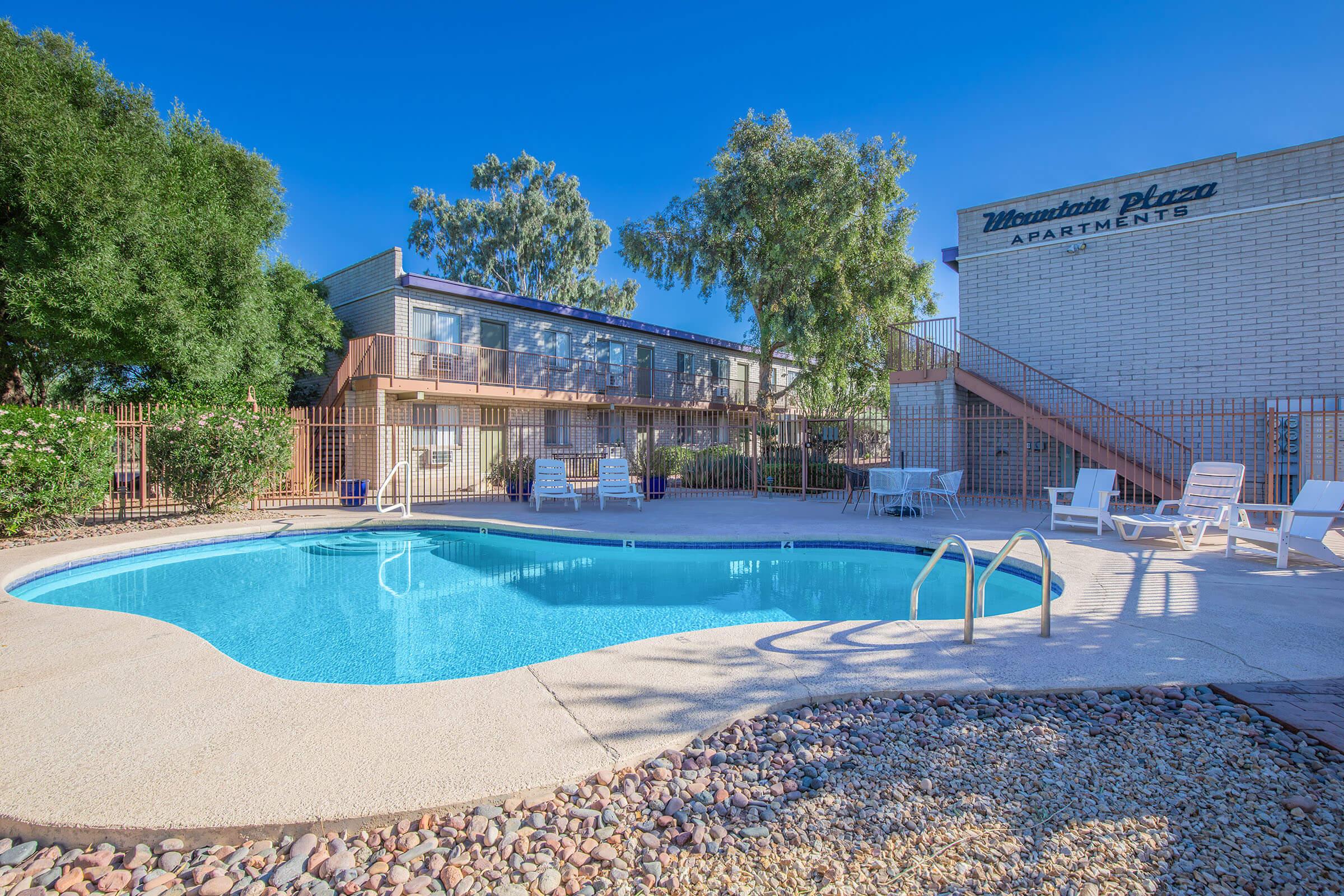 A clear blue swimming pool surrounded by lounge chairs, with a low-rise apartment building in the background. The scene is set under a bright blue sky, with trees providing some shade near the pool area. The building is labeled "Mountain Plaza Apartments."