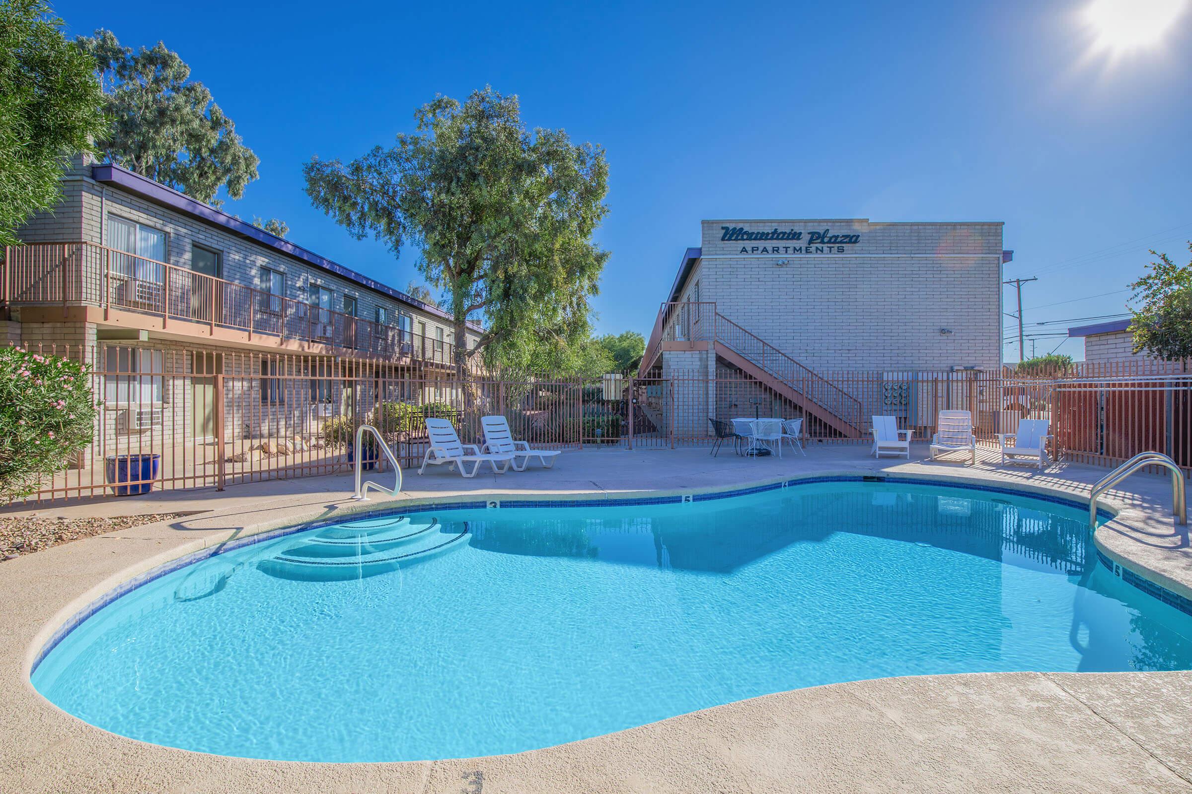 A serene outdoor pool area surrounded by apartment buildings. Sunlight reflects off the clear blue water, with lounge chairs positioned nearby. Lush trees provide some shade, creating a relaxing atmosphere for residents. The sign for "Mountain Plaza Apartments" is visible on a building in the background.