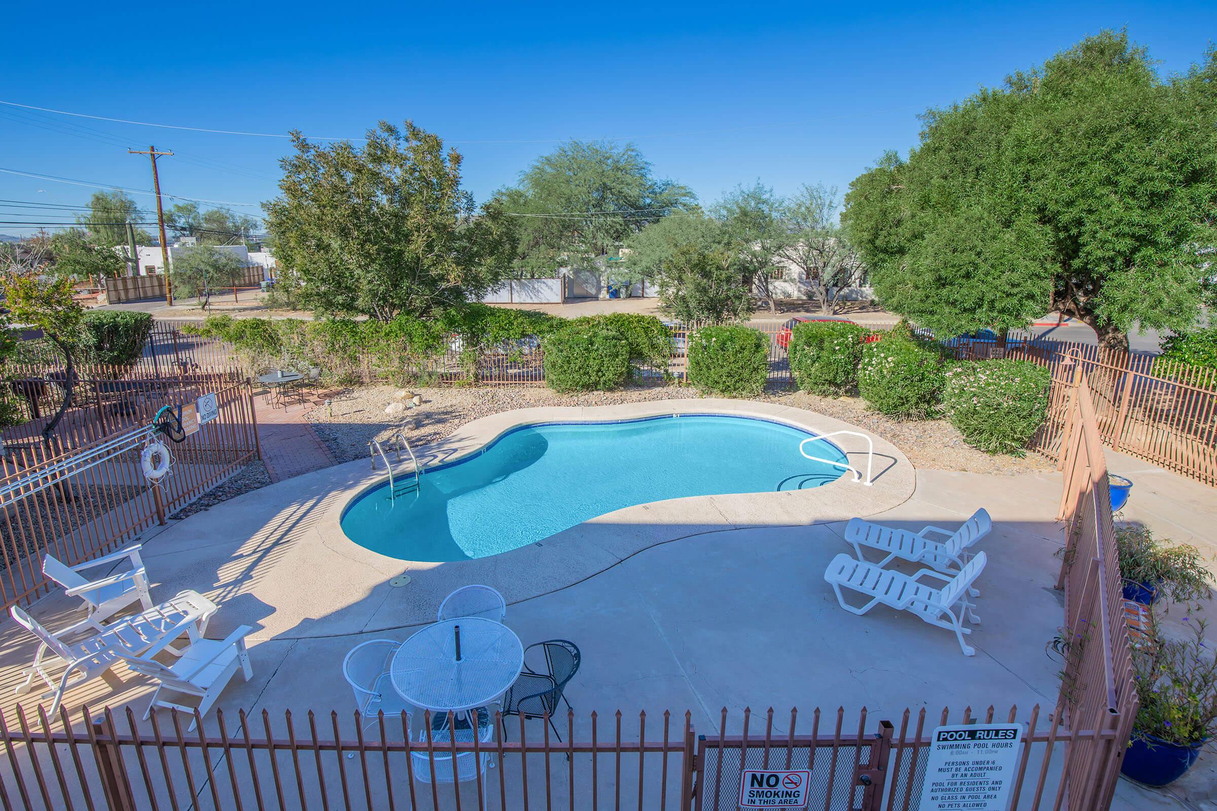 A view of a private swimming pool surrounded by a fence, with white lounge chairs and a small table nearby. Lush greenery is present in the background, and the sky is clear. The area is well-maintained and inviting, ideal for relaxation and leisure.