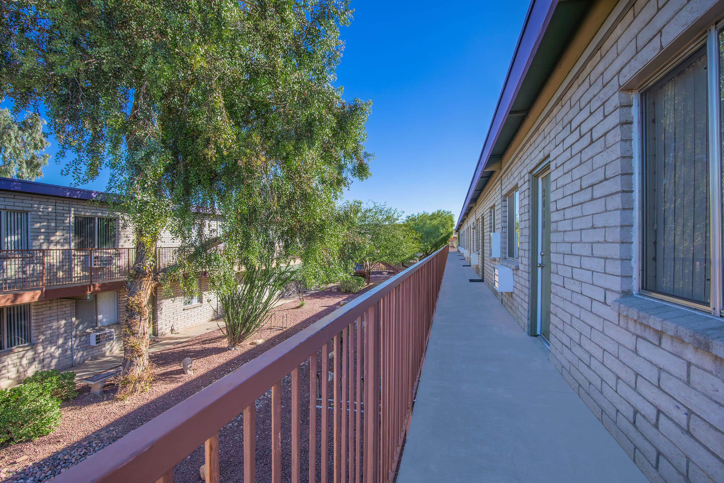 View from a balcony overlooking an apartment complex. The scene features a clear blue sky, green trees, and well-maintained landscaping with desert plants. The building has a simple brick facade and metal railings along the walkway, creating a serene residential atmosphere.