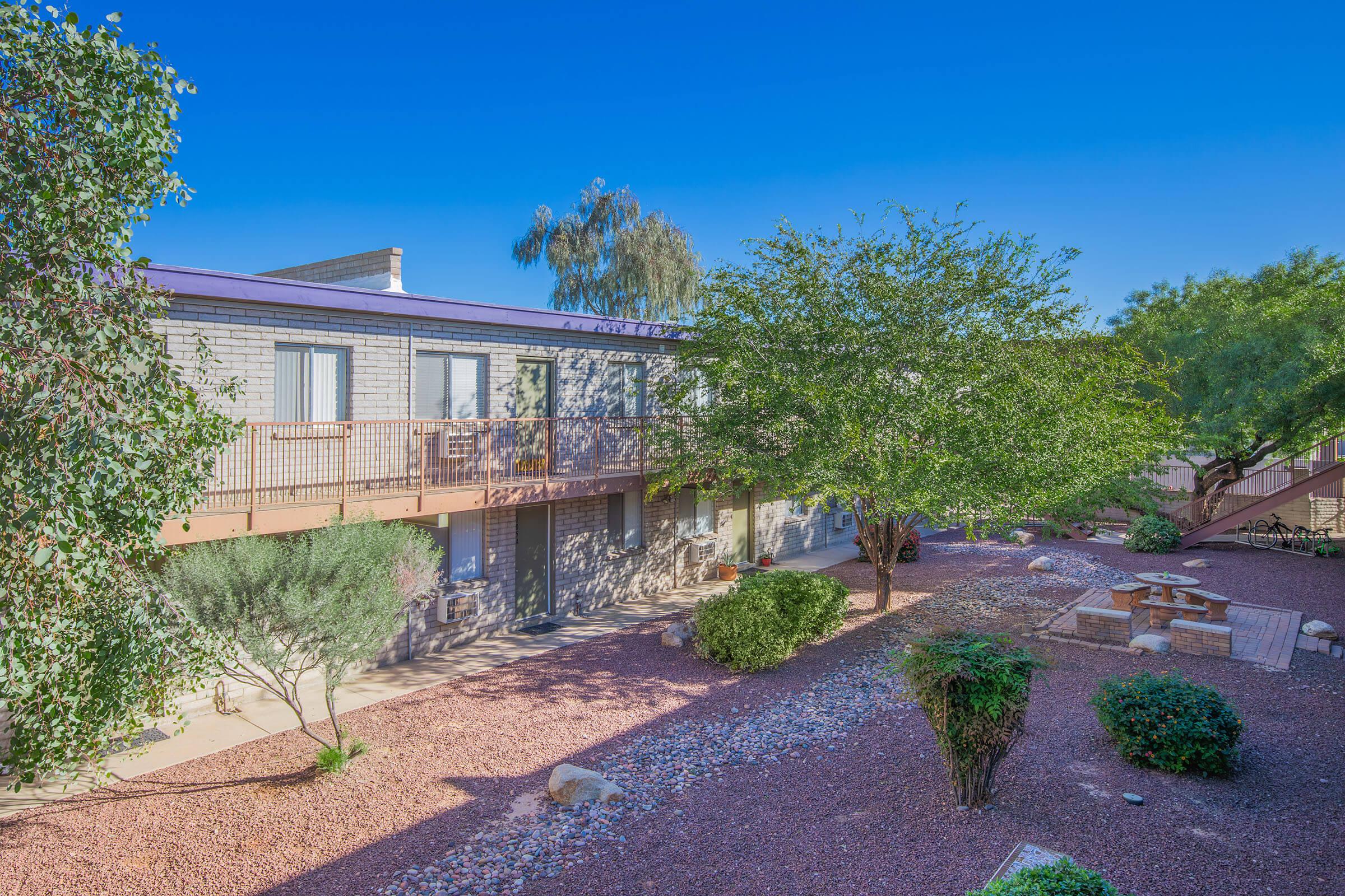 A view of a courtyard area with green shrubs and gravel, featuring a cozy seating arrangement and fire pit. Surrounding the courtyard are two-story buildings with balconies, under a clear blue sky. Decorative plants and desert landscaping enhance the outdoor space.