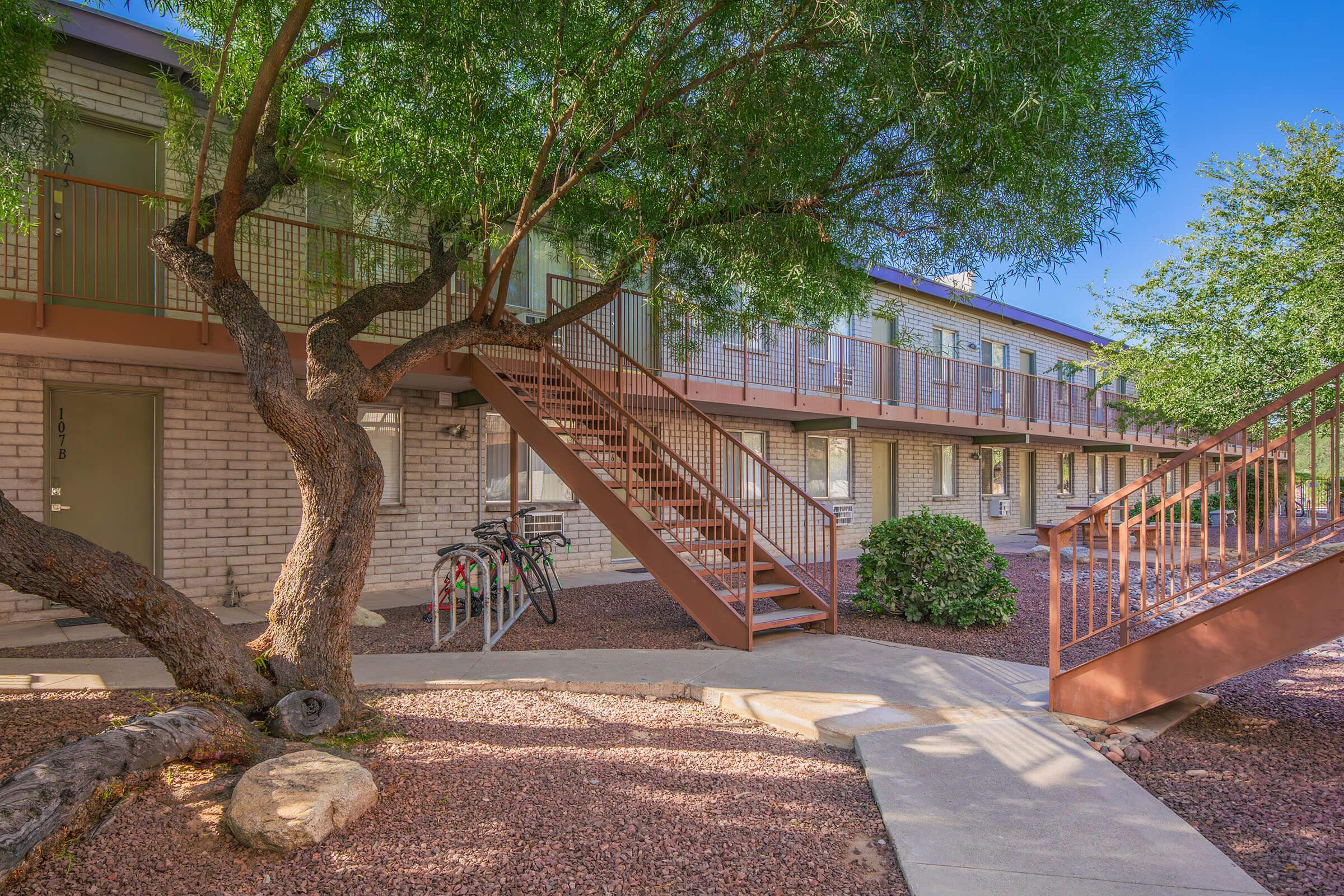 Exterior view of a two-story apartment building surrounded by trees. A staircase leads to the upper level, and there are bicycles parked on the side. The landscaping includes gravel and vegetation, creating a welcoming atmosphere. The architecture features a mix of brick and metal railings.