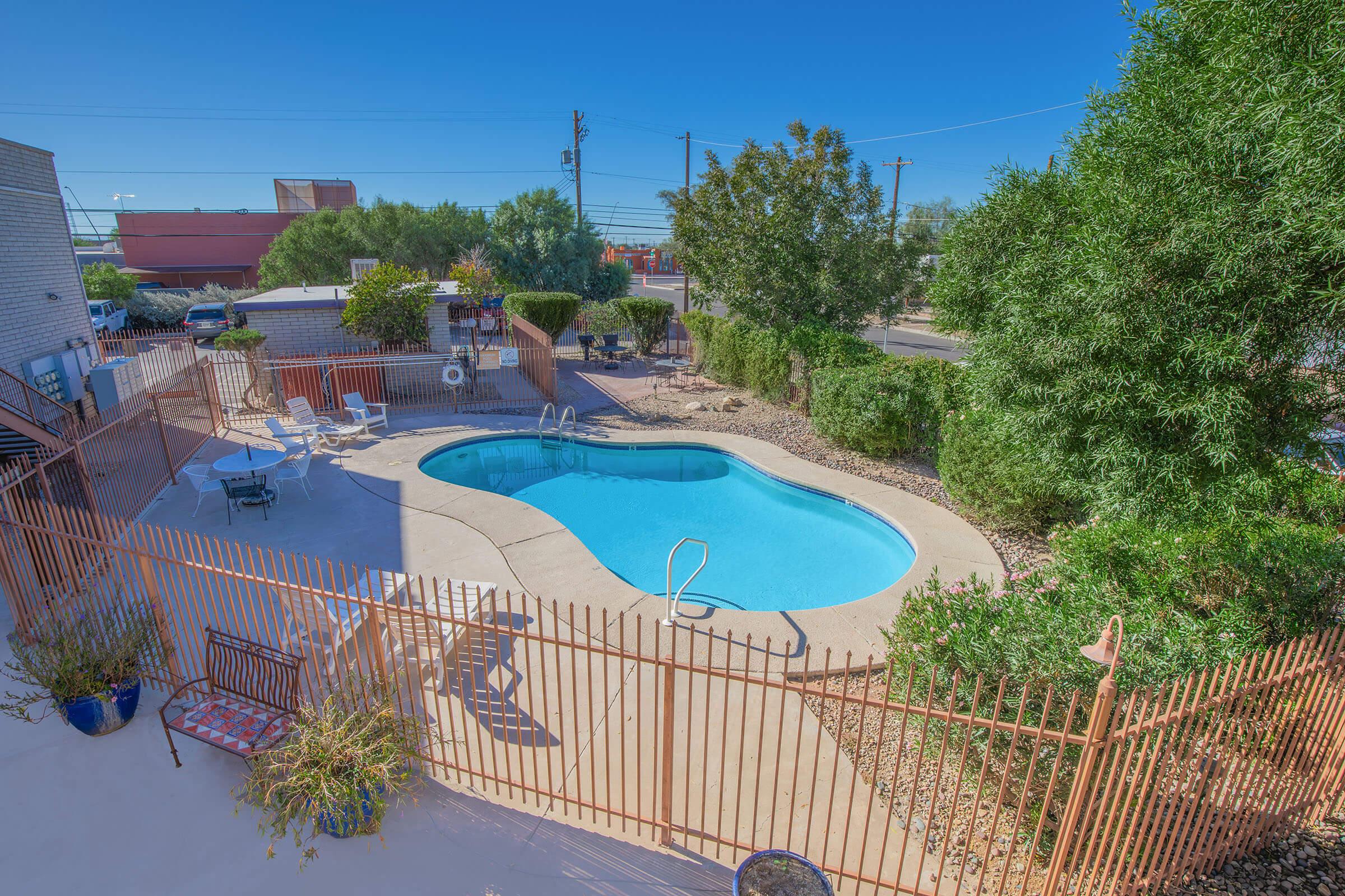 A scenic view of a swimming pool surrounded by a fenced area, featuring several lounge chairs and landscaped greenery nearby. Clear blue skies and sunlight enhance the inviting atmosphere of the pool area.