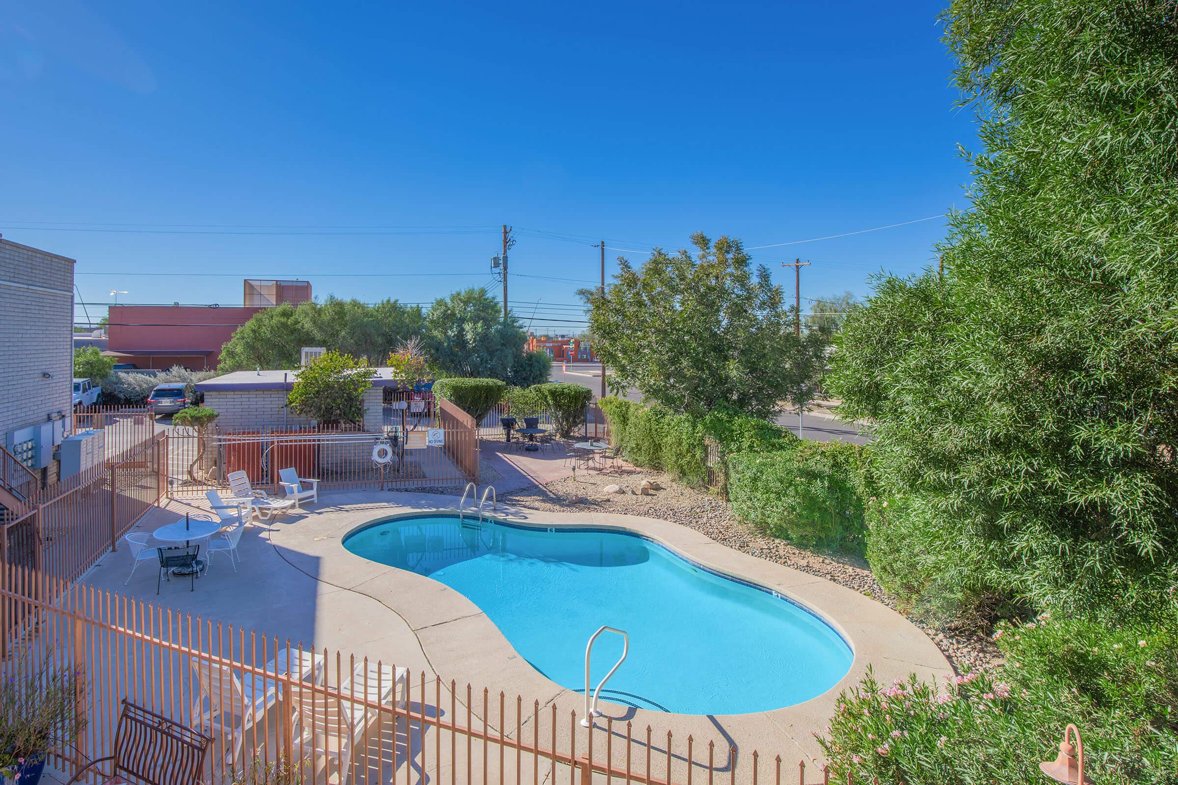 A clear blue swimming pool surrounded by a fence, with lounge chairs arranged nearby. Lush green trees line the area, and there are some buildings and power lines in the background under a bright blue sky. The setting appears inviting and well-maintained.