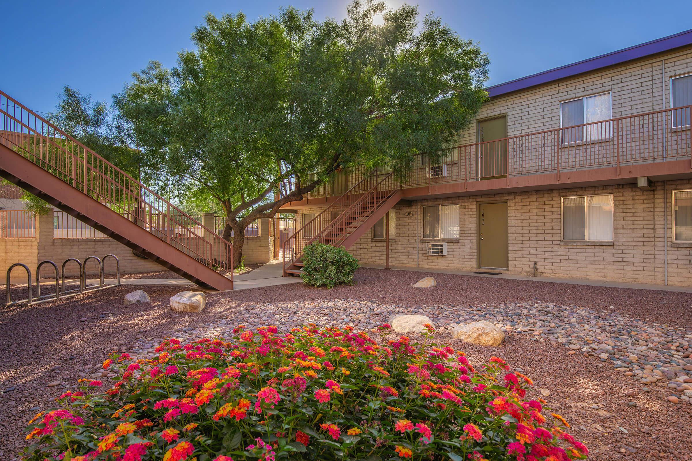 A view of an apartment complex surrounded by lush green trees and colorful flower beds. The building has two levels with a set of exterior stairs leading to the upper floor. There are also small stones and a gravel area in the foreground, enhancing the landscaping.