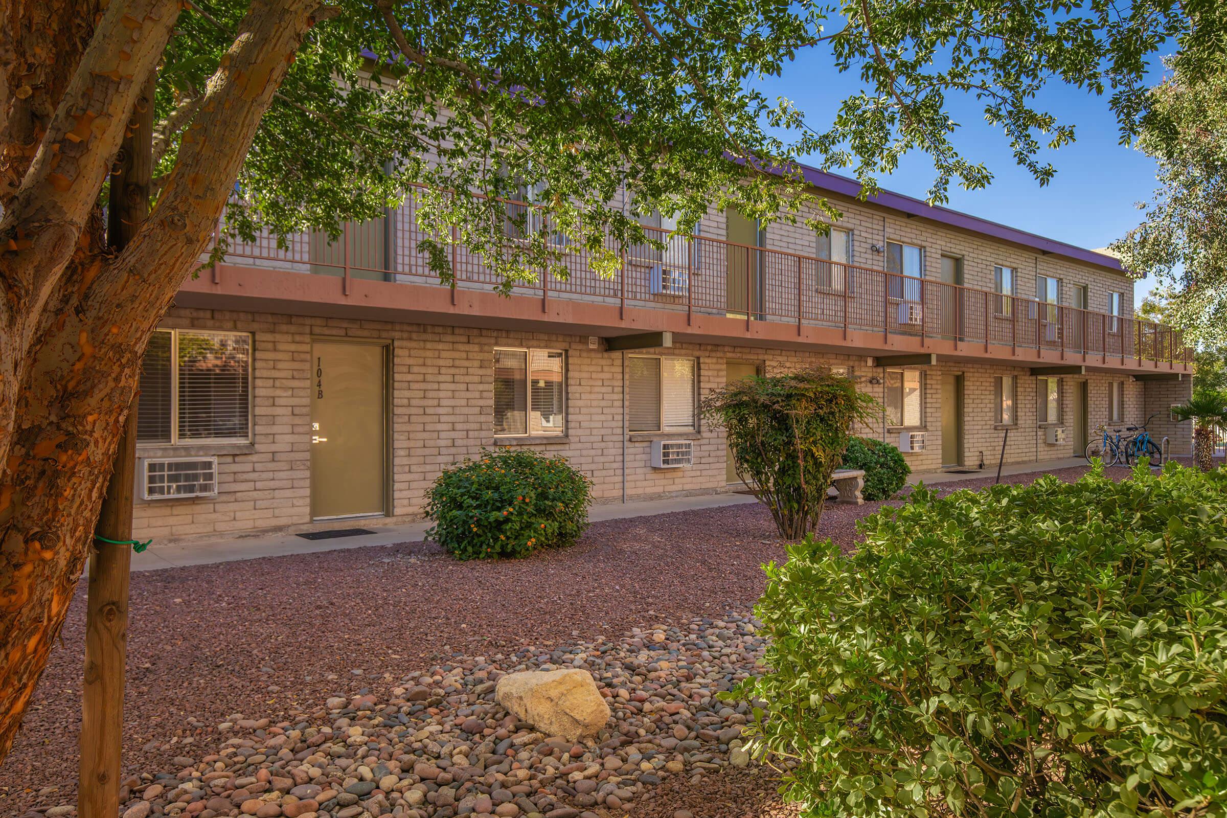 A view of a two-story brick apartment building surrounded by trees and shrubs. The building features multiple air conditioning units and a walkway leading to the doors. The landscape includes gravel pathways and rocks, creating a serene outdoor environment.