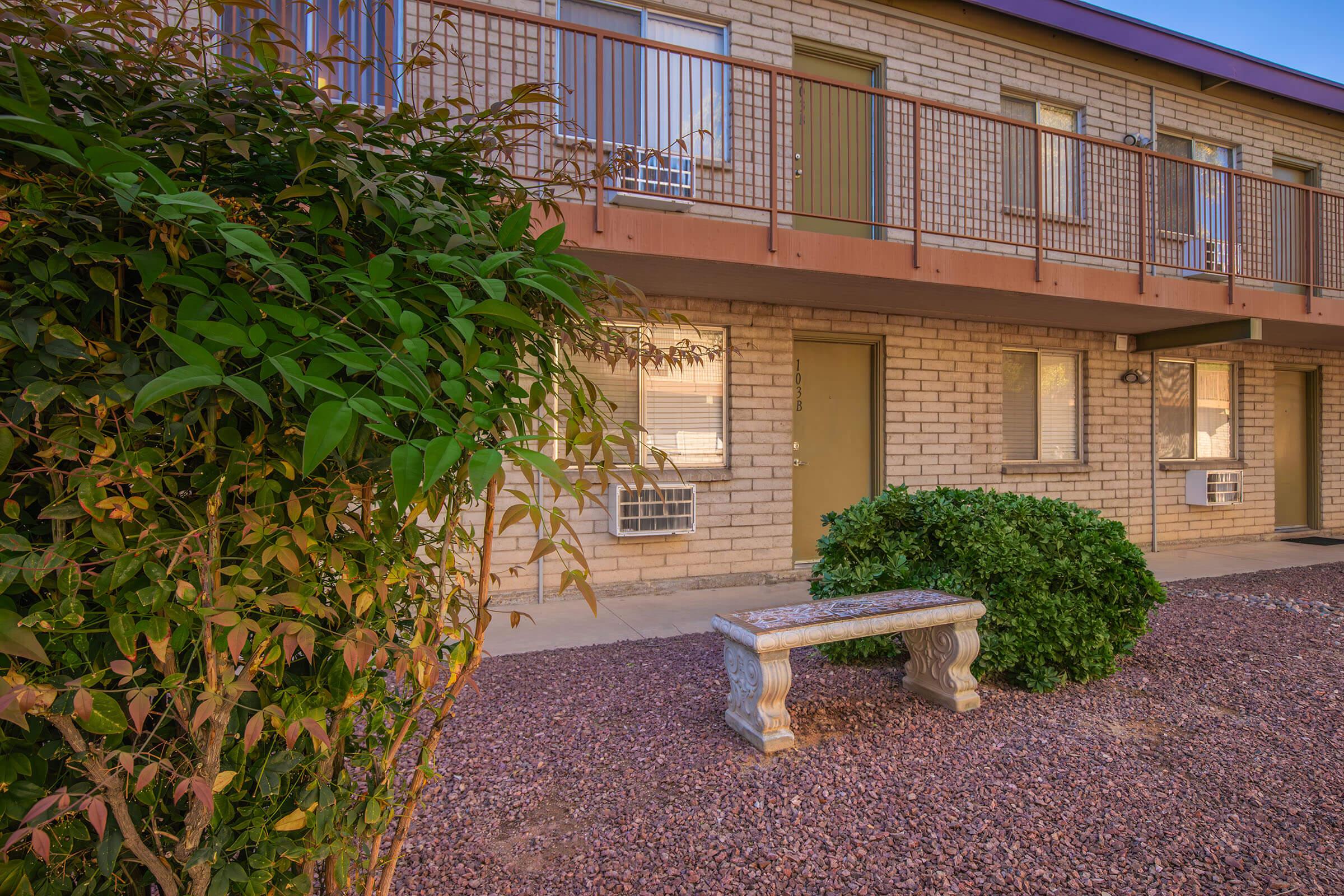 A brick building with a balcony and several windows. In front, there is a small, decorative stone bench and a bush. The ground is covered with gravel, and there are air conditioning units visible on the building's exterior. The setting appears to be well-maintained and inviting.