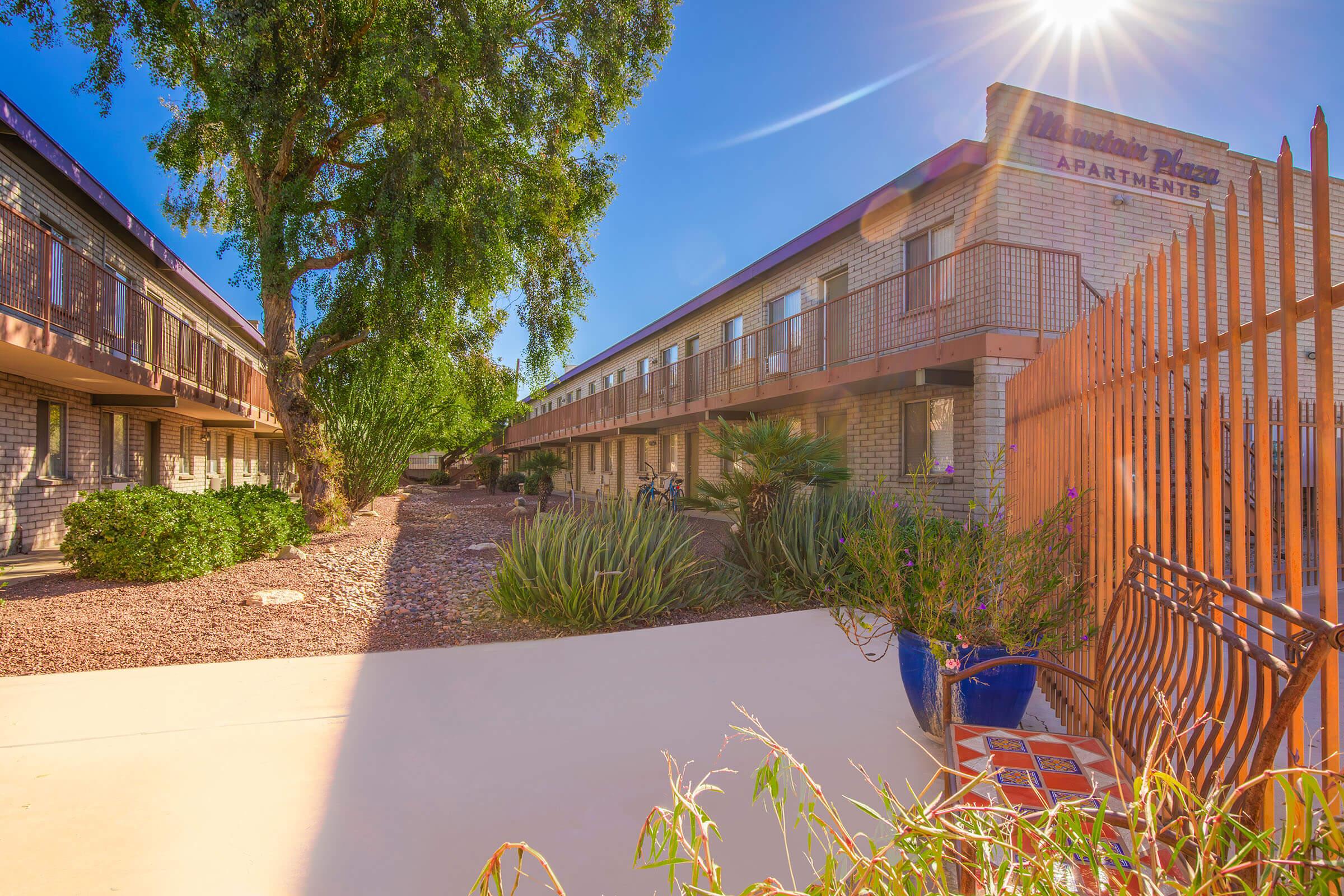 View of a multi-story apartment building labeled "Apartments" with a sunny blue sky overhead. The scene includes a pathway lined with plants and a decorative blue pot in the foreground. A wrought iron fence can be seen to the right, framing the entrance to the courtyard area.