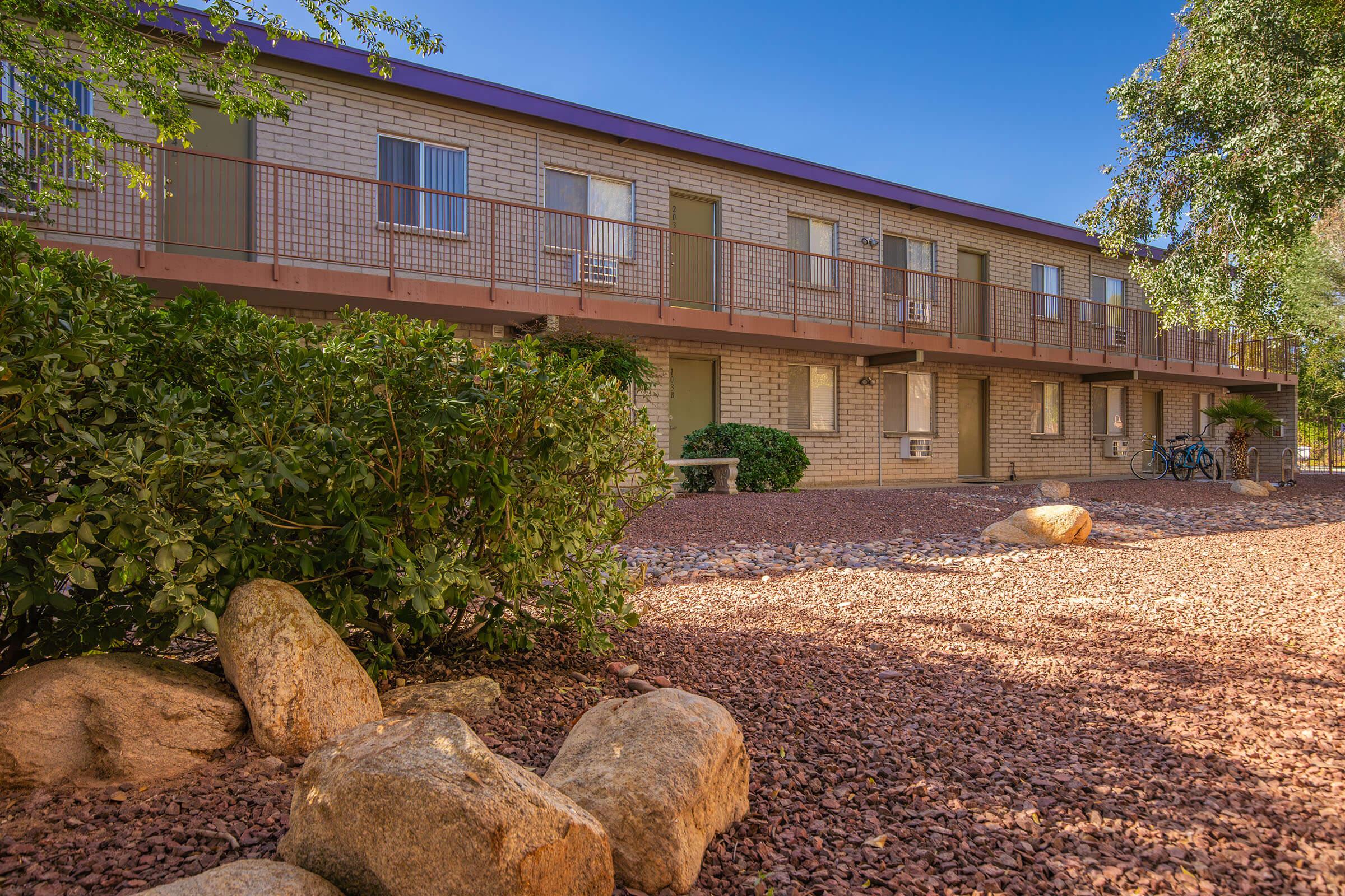 Two-story brick building with a purple roof and multiple windows, surrounded by gravel and bushy plants. The area features large rocks and bike racks in the background, with a clear blue sky overhead. The setting appears to be a well-maintained outdoor space.
