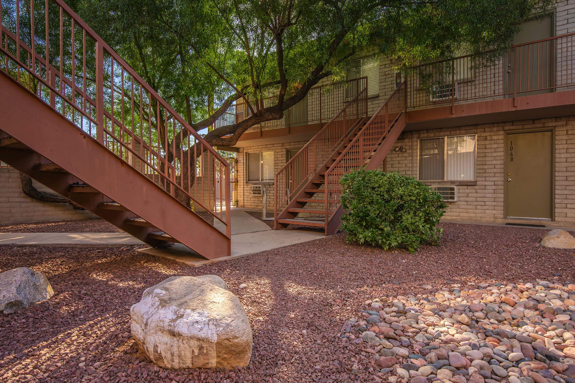 A view of an apartment complex courtyard featuring beige brick buildings. There are staircases with rust-colored railings leading to upper levels, surrounded by flowering shrubs and decorative rock landscaping. Sunlight filters through the foliage of nearby trees, creating a serene atmosphere.