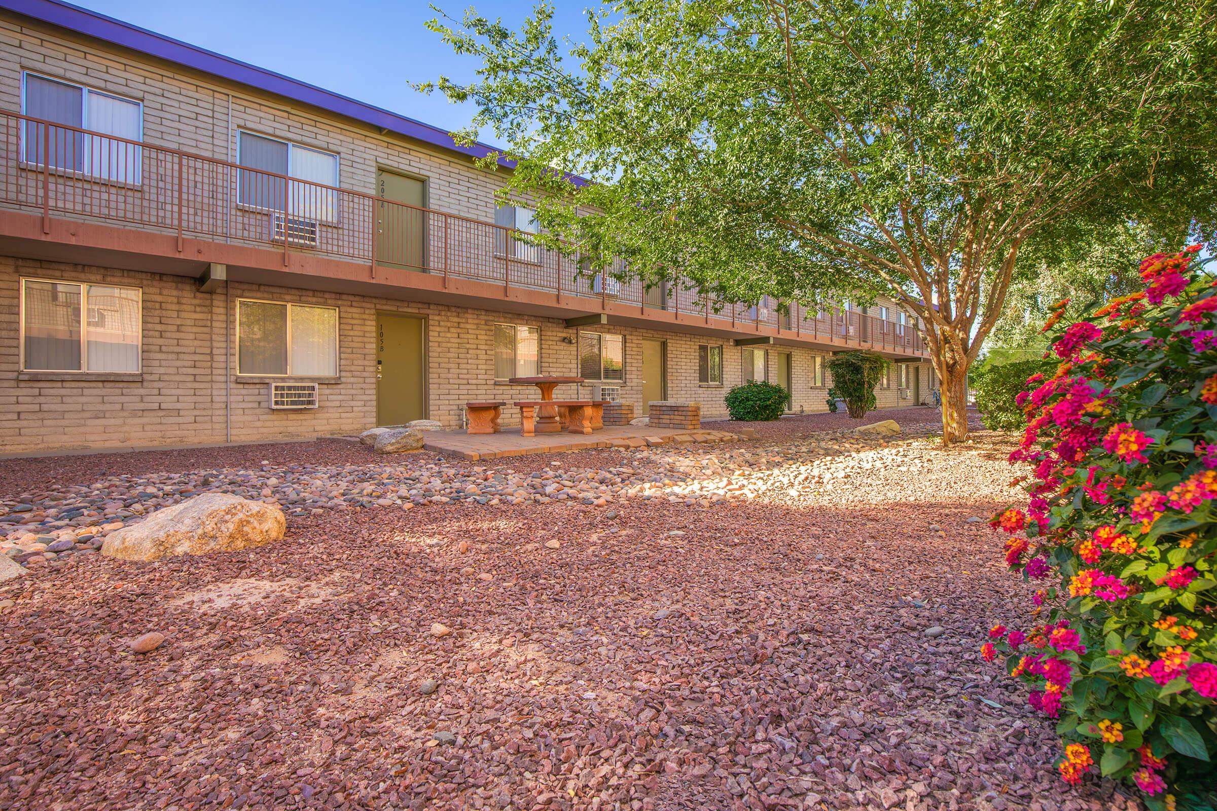 A tranquil scene featuring a low-rise building with a courtyard. The building has multiple windows and a green door. In the foreground, there's a gravel area with a wooden picnic table surrounded by lush greenery and colorful flowers, creating a peaceful outdoor environment.