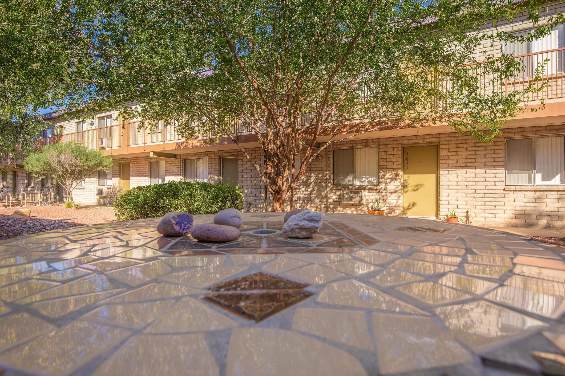 A close-up view of a decorative mosaic table featuring several smooth stones, with a leafy tree in the background. The table is situated in a courtyard area surrounded by buildings. Sunlight casts warm shadows, creating a peaceful, inviting atmosphere.