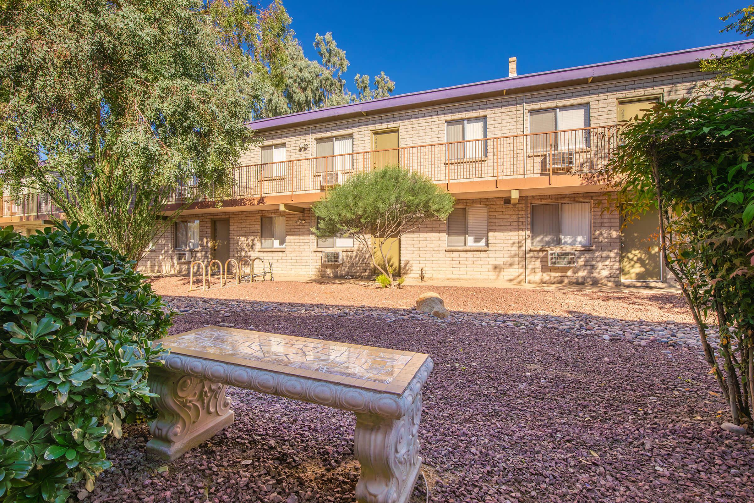 A view of a multi-story building surrounded by greenery and a gravel landscaping. In the foreground, a decorative stone bench sits nestled among low shrubs. The background features a well-maintained courtyard with bike racks and a sunny sky.