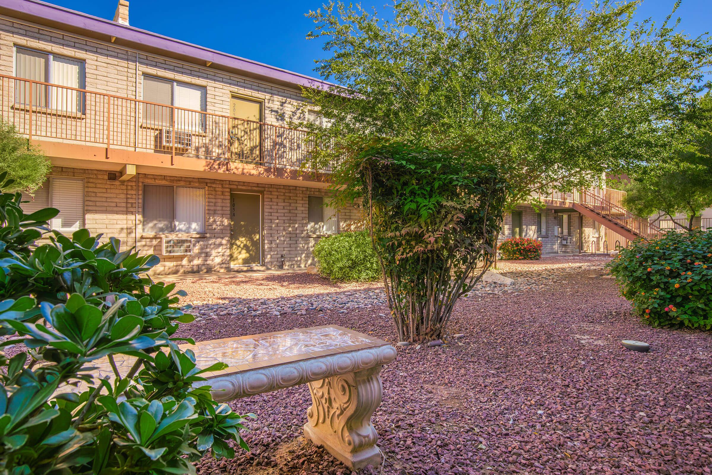 A landscaped courtyard featuring a stone bench surrounded by decorative rocks and lush green plants. In the background, two-story apartment buildings with balconies are visible under a clear blue sky. The scene conveys a serene, inviting atmosphere.