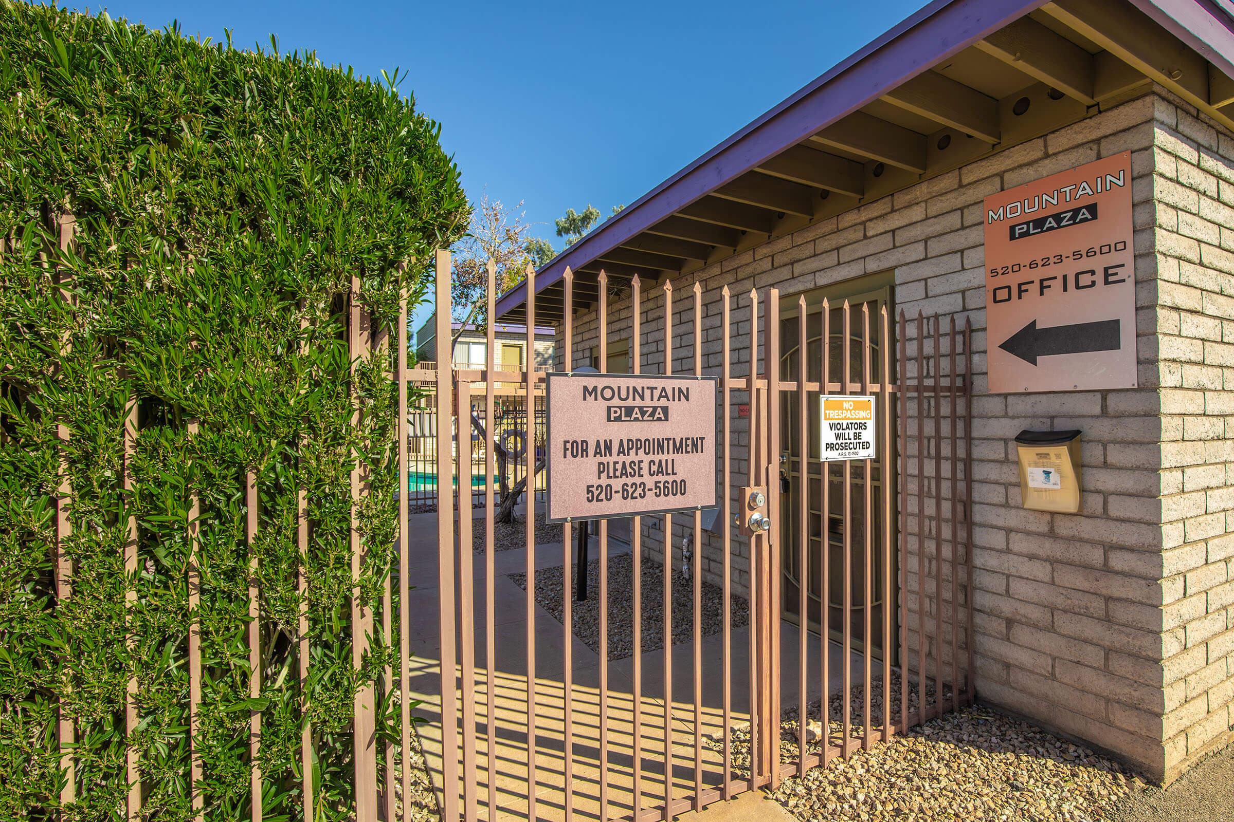 Gated entrance to Mountain Plaza office with signs for appointments, displaying the phone number 520-623-5600. The area features a well-maintained hedge, a pathway leading to the office, and a clear blue sky in the background.