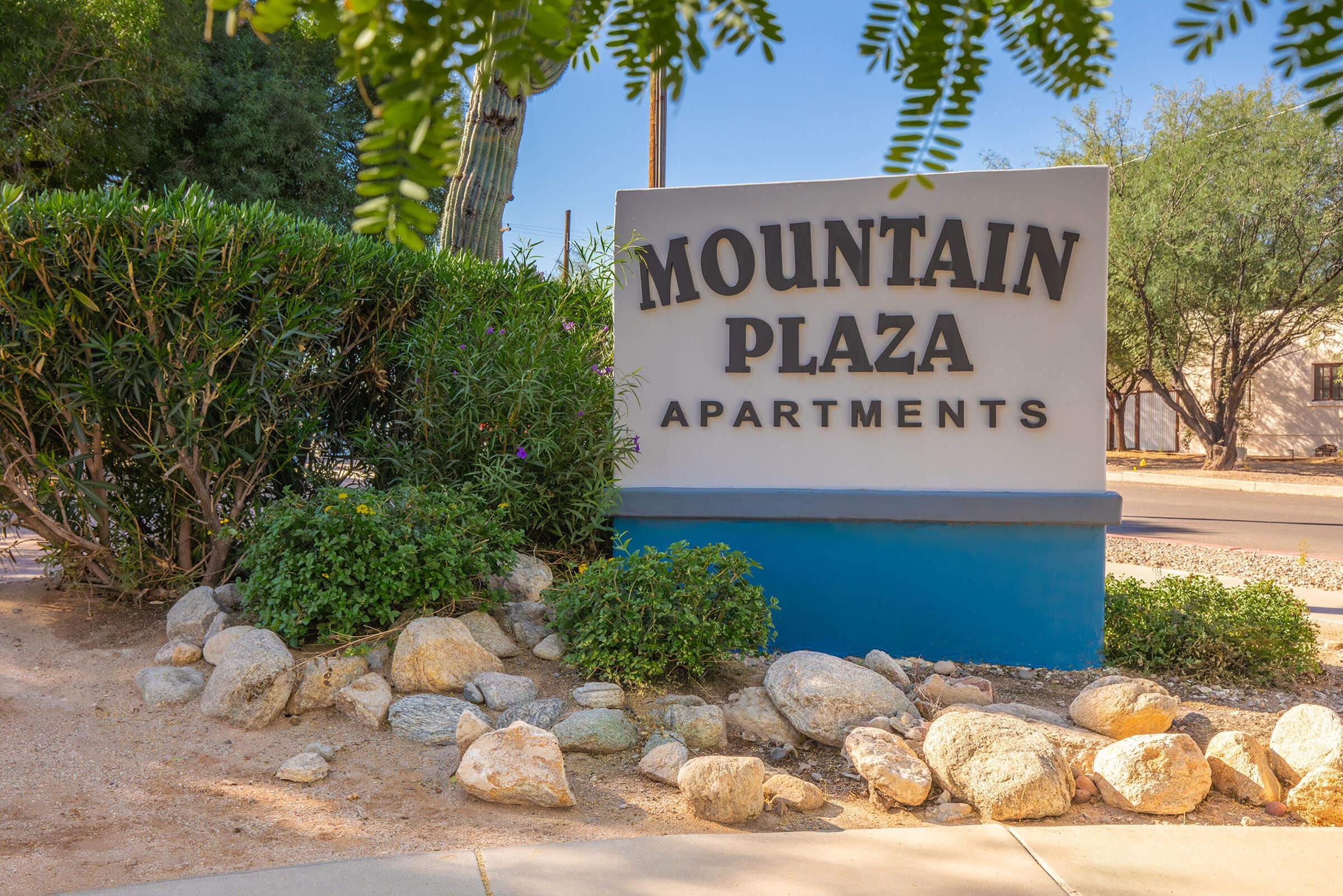 Sign for Mountain Plaza Apartments, prominently displayed in a landscaped area with rocks and greenery, set against a clear blue sky. The sign features a bold, modern font and indicates the name of the apartment complex.